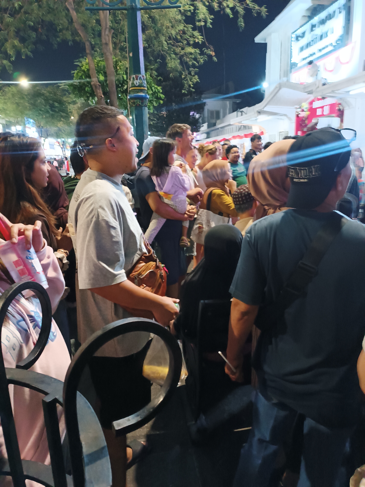 The image shows a crowd of people at night on a street in Yogyakarta, Indonesia. The street appears to be a busy thoroughfare with buildings lining one side. There are trees and streetlights visible. In the foreground, there are some black metal chairs. People are standing and walking, some are holding drinks, and some are carrying bags. There is a woman in a pink top and another woman with long brown hair. A man in a grey t-shirt is in the center, with a brown bag slung over his shoulder. A woman is holding a baby. The overall impression is of a lively urban scene at night.