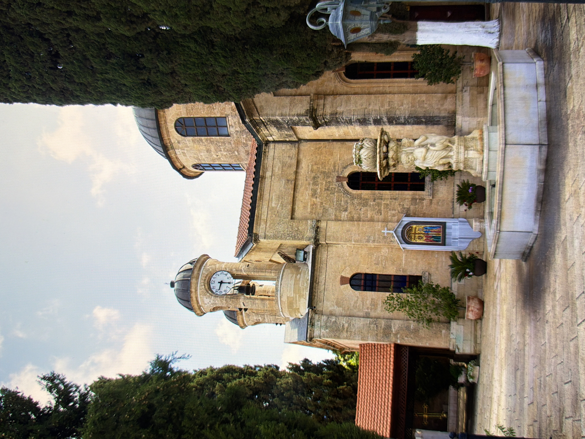 This media file shows a tall, light-colored stone building with a domed roof and arched windows. The building appears to be a church or a similar religious structure. A prominent feature is a clock tower with a round clock face displaying Roman numerals. In front of the building, there's a stone fountain with sculpted figures. The surrounding area is filled with lush green trees, and the sky is partly cloudy, suggesting daytime. The scene is shot from a low angle, looking up at the building.
The image seems to be a still photograph of the exterior of the building. There are no people visible in the frame, and no specific activity or event is taking place. The lighting suggests it's daytime, possibly mid-morning or afternoon, and the weather appears fair. The architecture of the building, with its stone construction and decorative elements like the fountain and stained glass window, suggests a historical or significant location. While the location context is provided as Kyiv, Ukraine, there are no explicit visual cues within the image itself that definitively confirm this specific location. No visible text is present in the image.