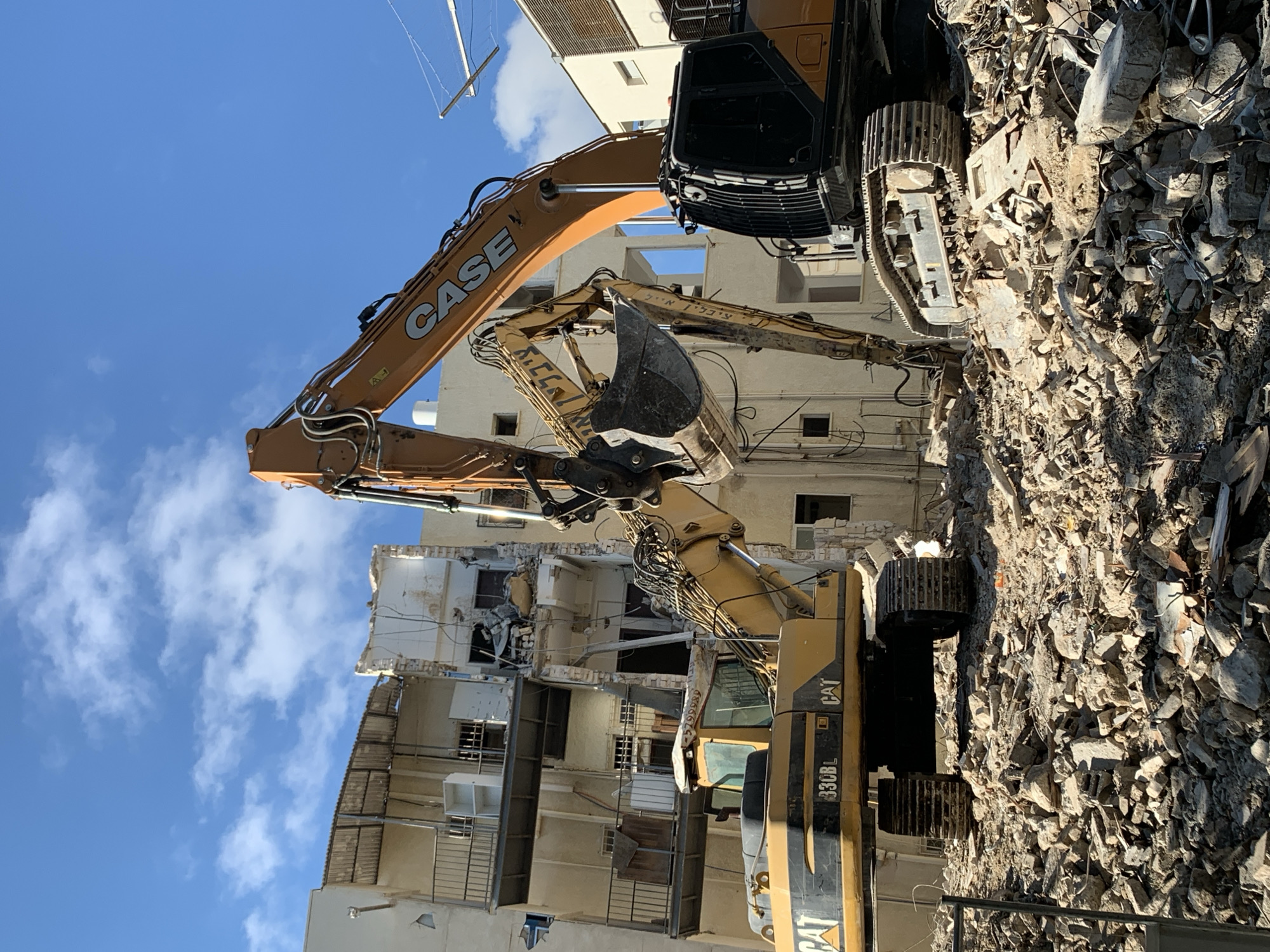 The image shows a construction site in Tel-Aviv, Israel, where a building is being demolished. Two excavators, one a Case and the other a CAT 330BL, are actively demolishing the structure. The debris of the demolition is piled up around the excavators. The sky is blue with some clouds.
