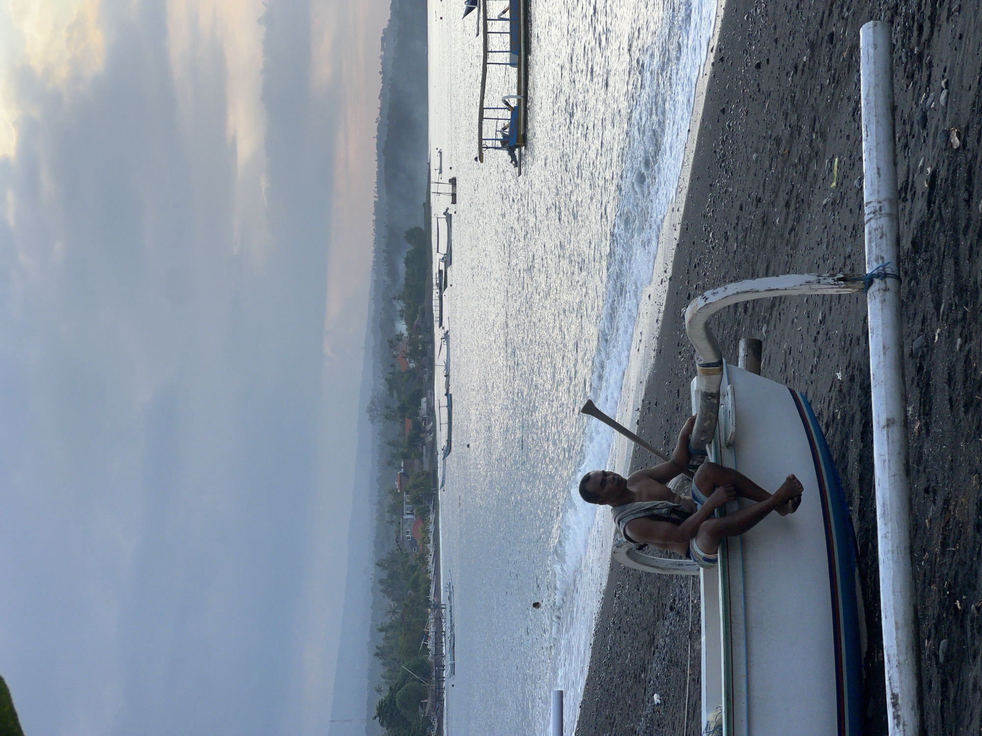 In the city of Abang, Indonesia, a man sits on the edge of a beached outrigger canoe on a dark, pebbly shore. The canoe is white with blue and red stripes along its hull. The man, shirtless and wearing striped shorts, has a towel draped over his left shoulder and is holding a wooden pole. The ocean waves gently lap the shore, and several other boats are visible on the water. In the background, a hazy coastline is lined with lush greenery and scattered buildings. The sky is overcast, with soft clouds tinged by the fading light.