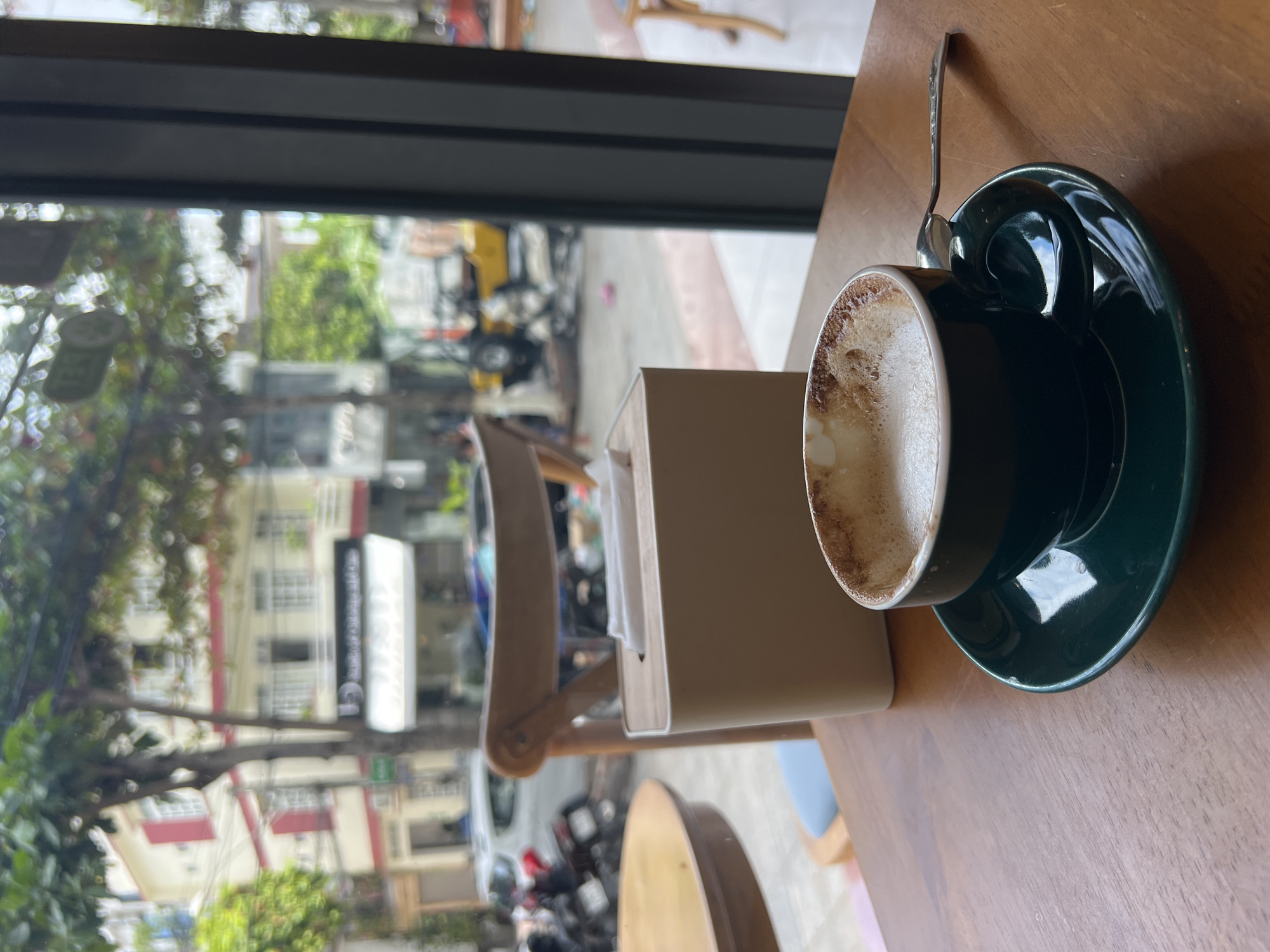Captured from an indoor perspective, the image presents a coffee cup on a wooden table, positioned adjacent to a window offering a view of the urban landscape. The coffee cup, dark green in color and resting on a matching saucer, sits next to a tissue holder. A small spoon lies beside the cup. The window reveals a street scene with buildings and trees, and an urban environment suggesting a city or town setting. The street is filled with parked motorcycles. The urban environment and street scene, with the buildings and motorbikes, suggest the city of Nha Trang Ward, Vietnam.