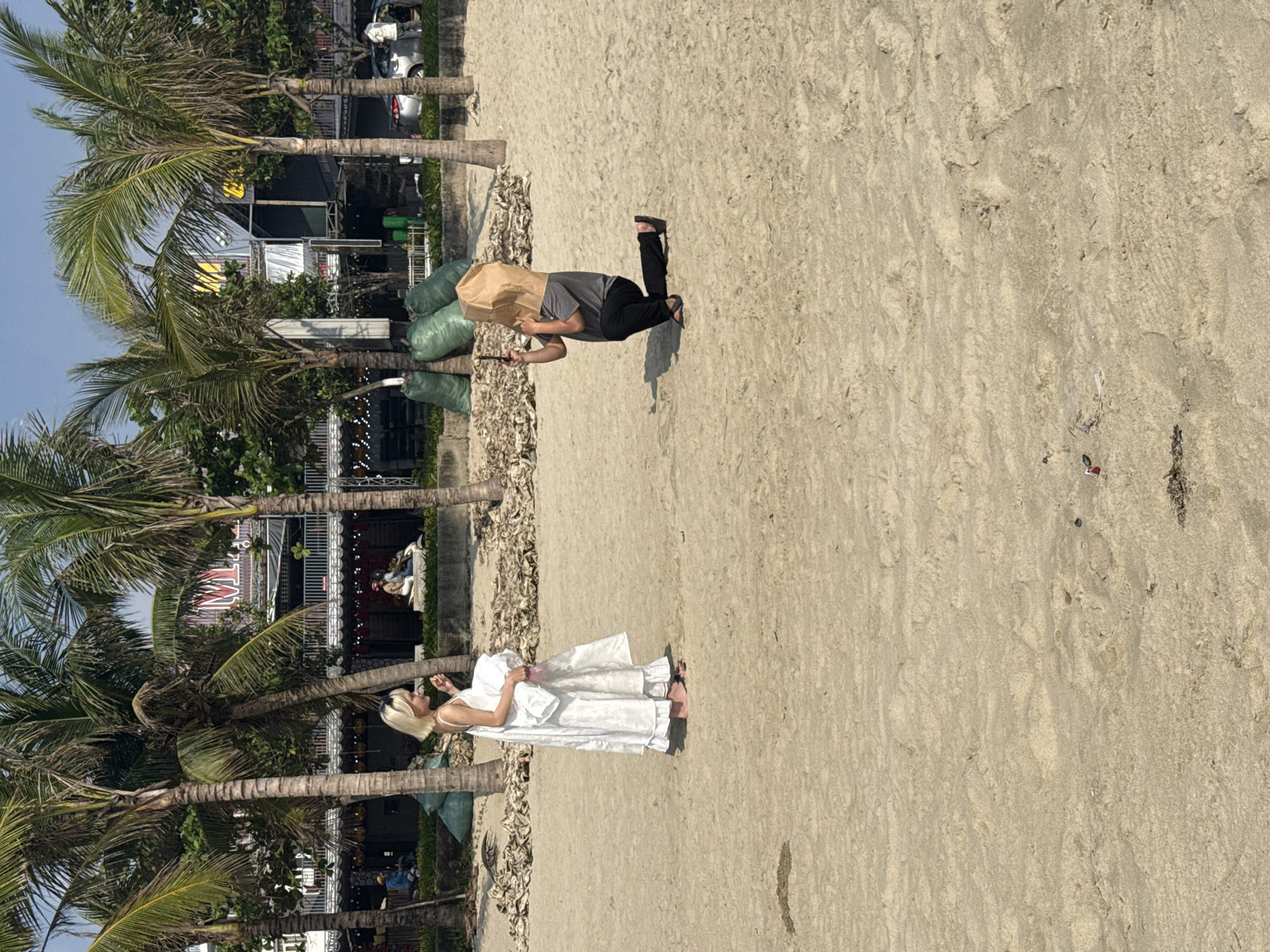 A horizontal daytime shot depicts a beach scene. The beach has light tan sand. Two individuals are present. A woman in a long white dress stands near a palm tree. A man wears dark pants, a gray shirt, and a brown paper bag over his head. He is holding a slim black object. Several large green bags are near him. Palm trees line the edge of the beach near buildings. Đà Nẵng, Vietnam is implied.