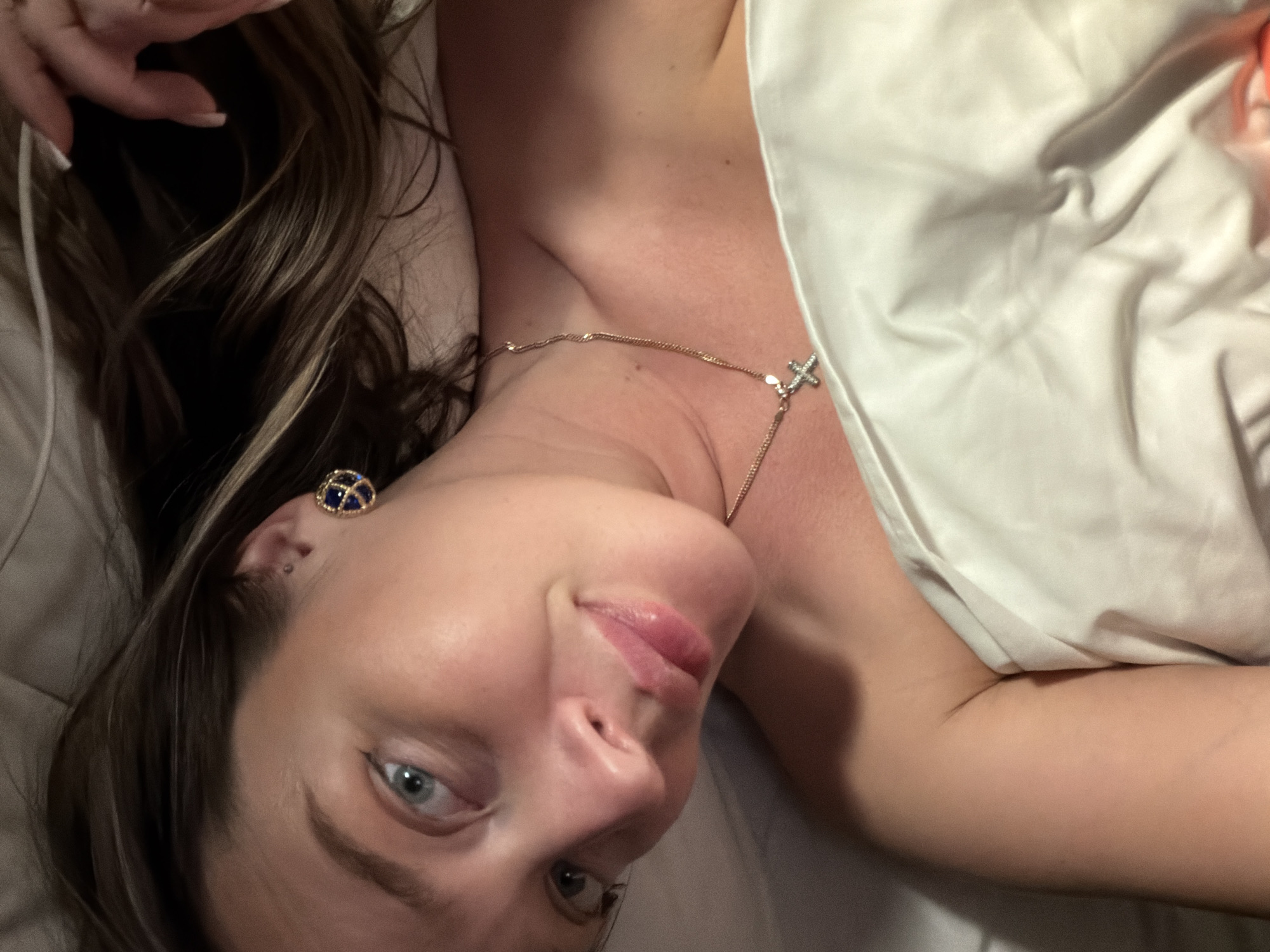 The image shows a woman lying in bed. She has light skin, brown hair with blonde highlights, and blue eyes. She is wearing a gold necklace with a cross pendant and a dark blue earring. She is partially covered by a white blanket. The background is blurred and indistinct. The location is not identifiable as Moscow, Russia.