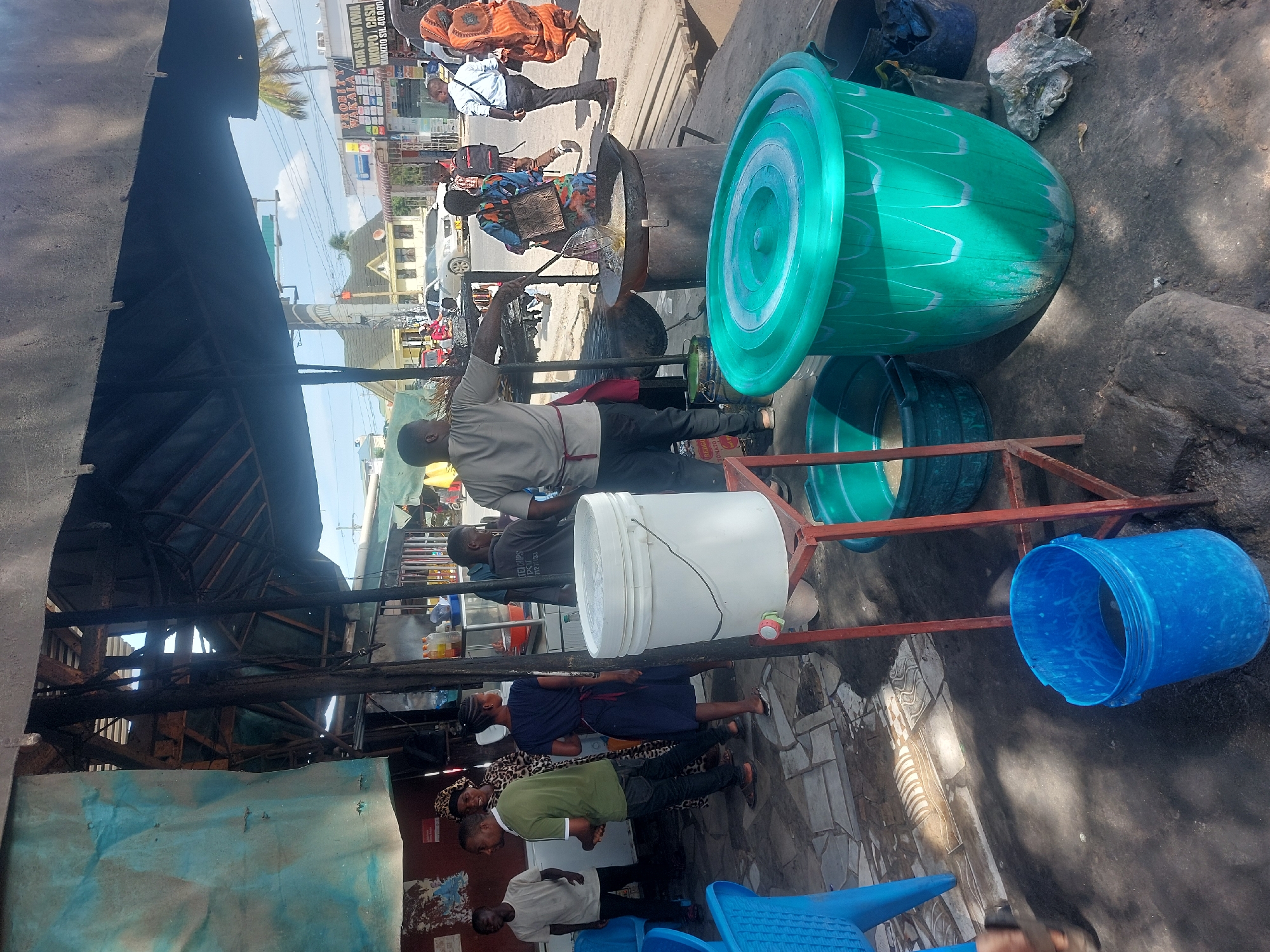 The image captures a bustling street scene in Dar es-Salaam, Tanzania. A man is actively involved in deep-frying food in a large pot, likely preparing street food for sale. There are several people in the vicinity, some of whom appear to be customers or passersby. A stack of plastic containers in green, white, and blue hues is present, possibly used for storage or transportation. A blue plastic chair is also in view. The overall atmosphere is that of a busy commercial area, typical of Dar es-Salaam, with various structures and signs visible in the background.