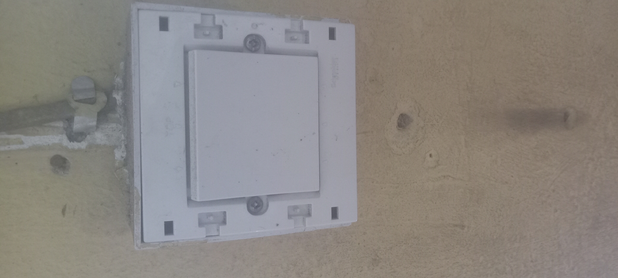 The image shows a white electrical switch plate attached to a pale yellow wall in Mbribit, Nigeria. The switch plate is square with a rectangular switch in the center, secured by two visible screws. There are small square and T-shaped features around the edges of the plate. The wall shows signs of wear, including exposed areas of bare substrate, two drilled holes, and a metal conduit attached to the wall with a metal fastener above and to the left of the switch plate. The environment appears to be interior. No people or interactions are visible in the frame.