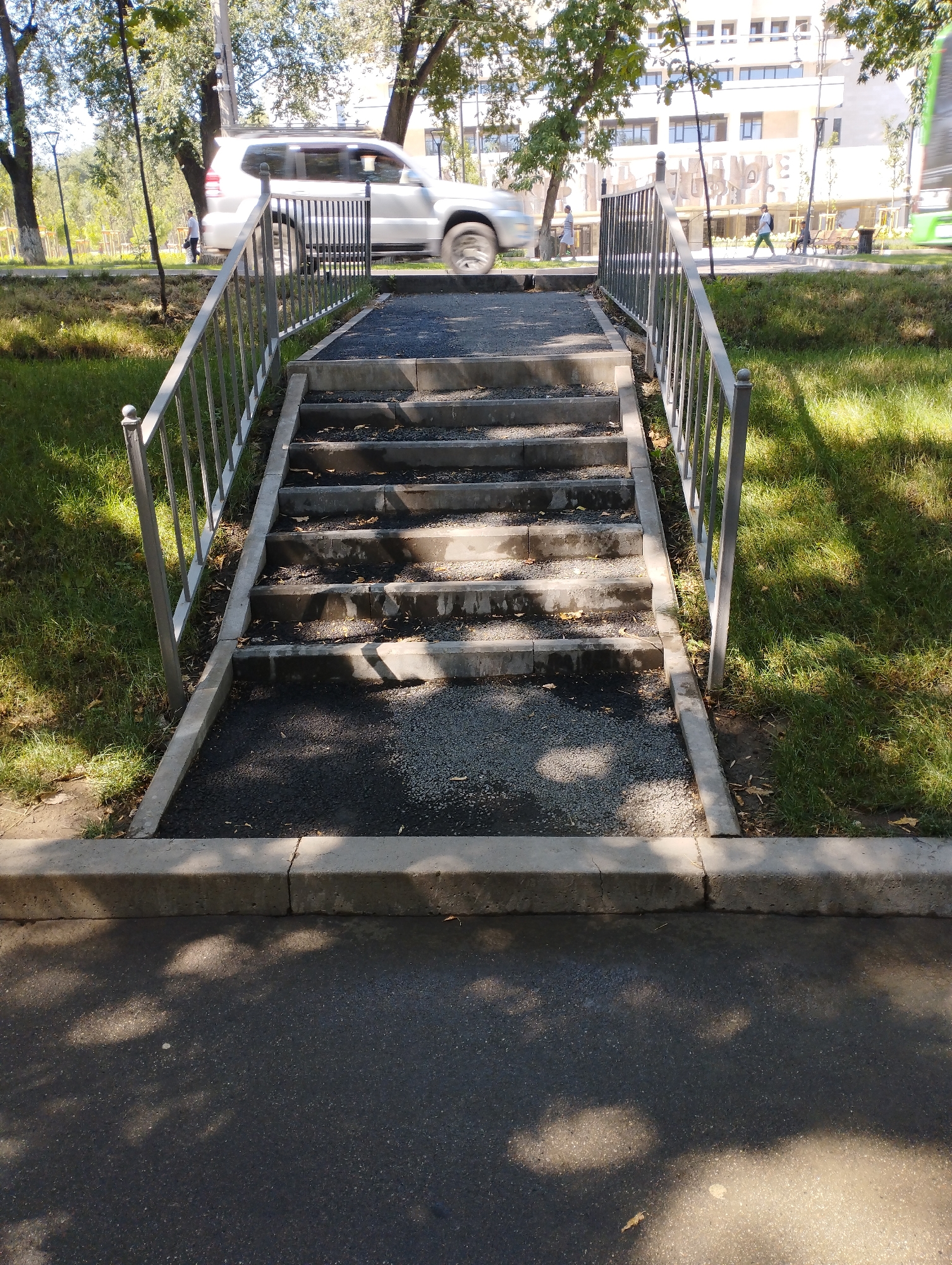 An outdoor, eye-level shot captures a set of concrete stairs with metal railings, leading upwards from a paved area. The stairs, covered with gravel and asphalt, are worn and have scattered leaves. To the left and right of the stairs, there is a strip of green grass. In the background, trees with green foliage are visible, along with a white SUV in motion and a modern building. People are present on the sidewalk in the background. The scene appears to be a public space in Almaty, Kazakhstan, likely a park or a residential area, given the presence of the building and park-like landscaping. The lighting suggests it is daytime.