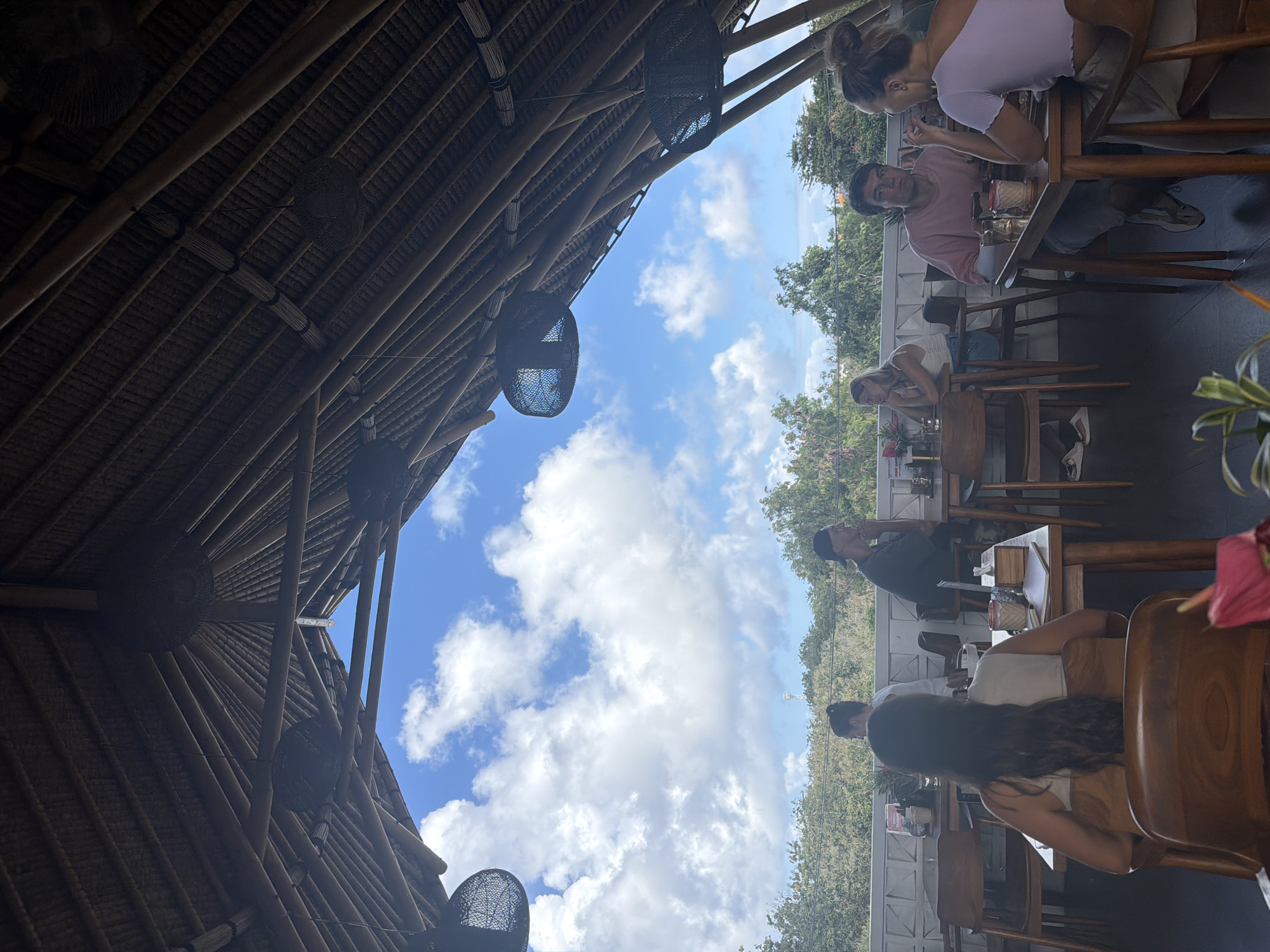 Captured indoors, a sunny day view under a bamboo structure with woven light fixtures in Kuta Selatan, Indonesia. Several patrons are seated at wooden tables and chairs. They are engaged in dining or conversation. The view beyond the structure shows green vegetation and a blue sky dotted with white clouds. The camera angle is looking upward.