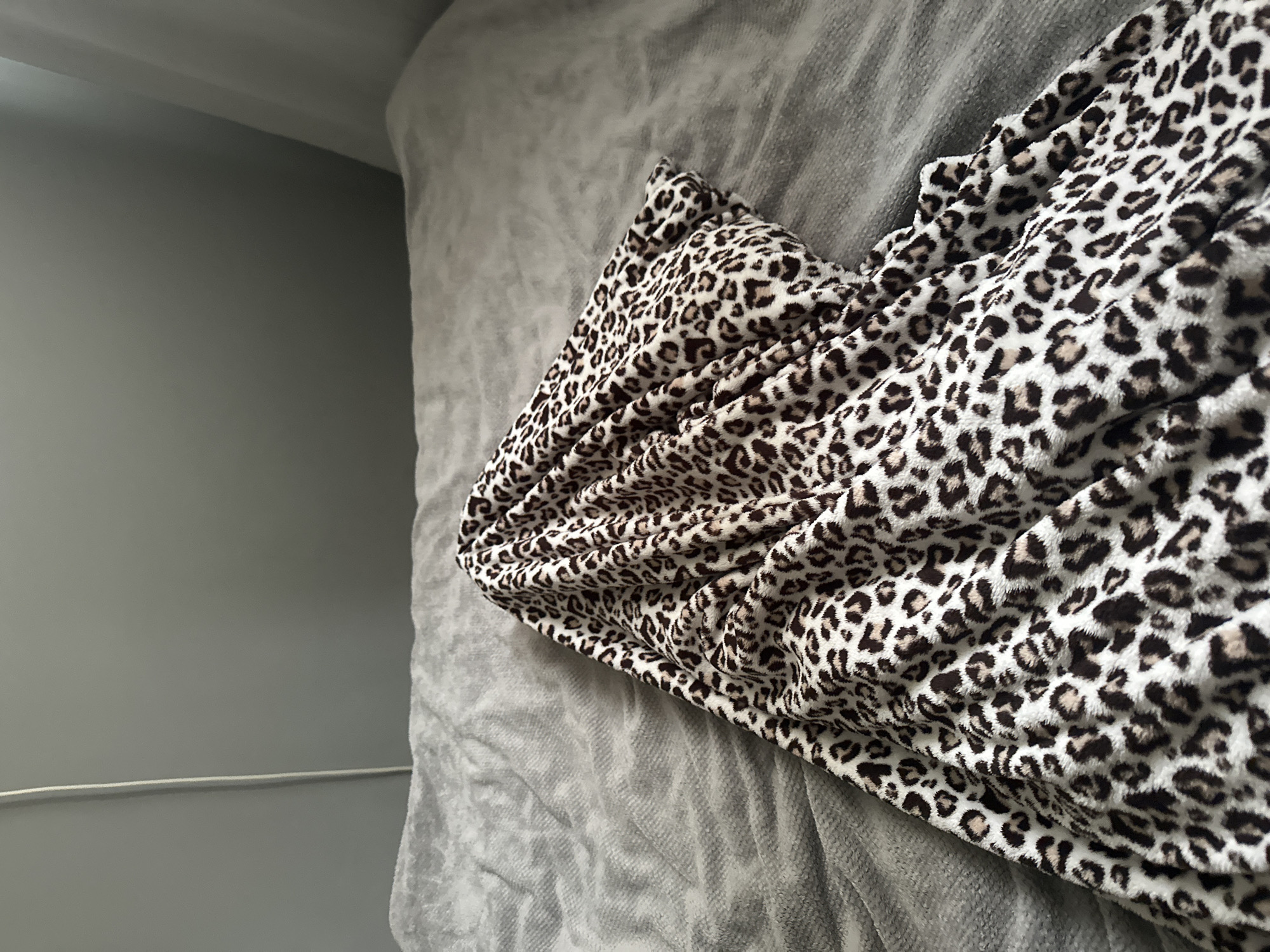 The image displays an indoor scene in Unknown, Unknown, focusing on textiles within a room. In the foreground and extending towards the right-center, a rumpled leopard print blanket or fabric, characterized by white, brown, and black irregular spots, is prominently featured. Adjacent to and partially underneath this fabric, a grey, textured blanket or bedspread covers a flat surface, showing multiple folds and creases. The upper background consists of a plain grey wall. A thin, vertical white cable is affixed to the grey wall on the upper left side of the frame. A portion of a white curtain is visible on the far right edge of the background. No individuals are present, and no discernible actions are occurring.