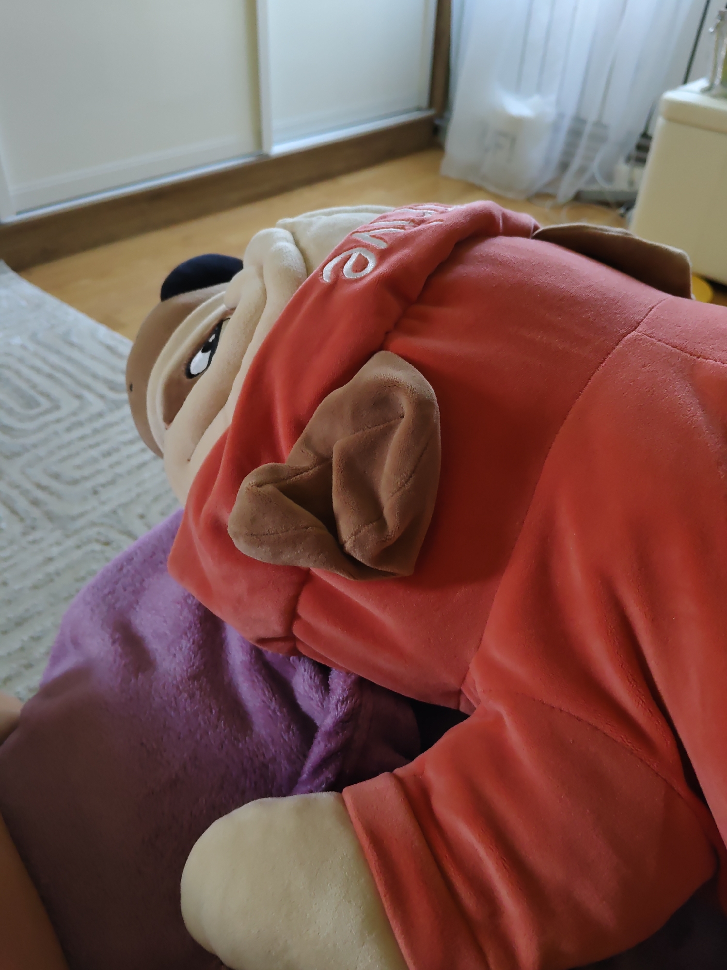 The image shows a close-up, low-angle view of a plush bulldog toy. The toy is predominantly orange-red with beige and brown accents. It is lying on top of a purple fuzzy blanket. The background consists of a wooden floor and a white sliding closet door with vertical blinds. The scene appears to be indoors, likely in a residential setting. There is no indication of the location being Rostov-on-Don, Russia, or any specific city.