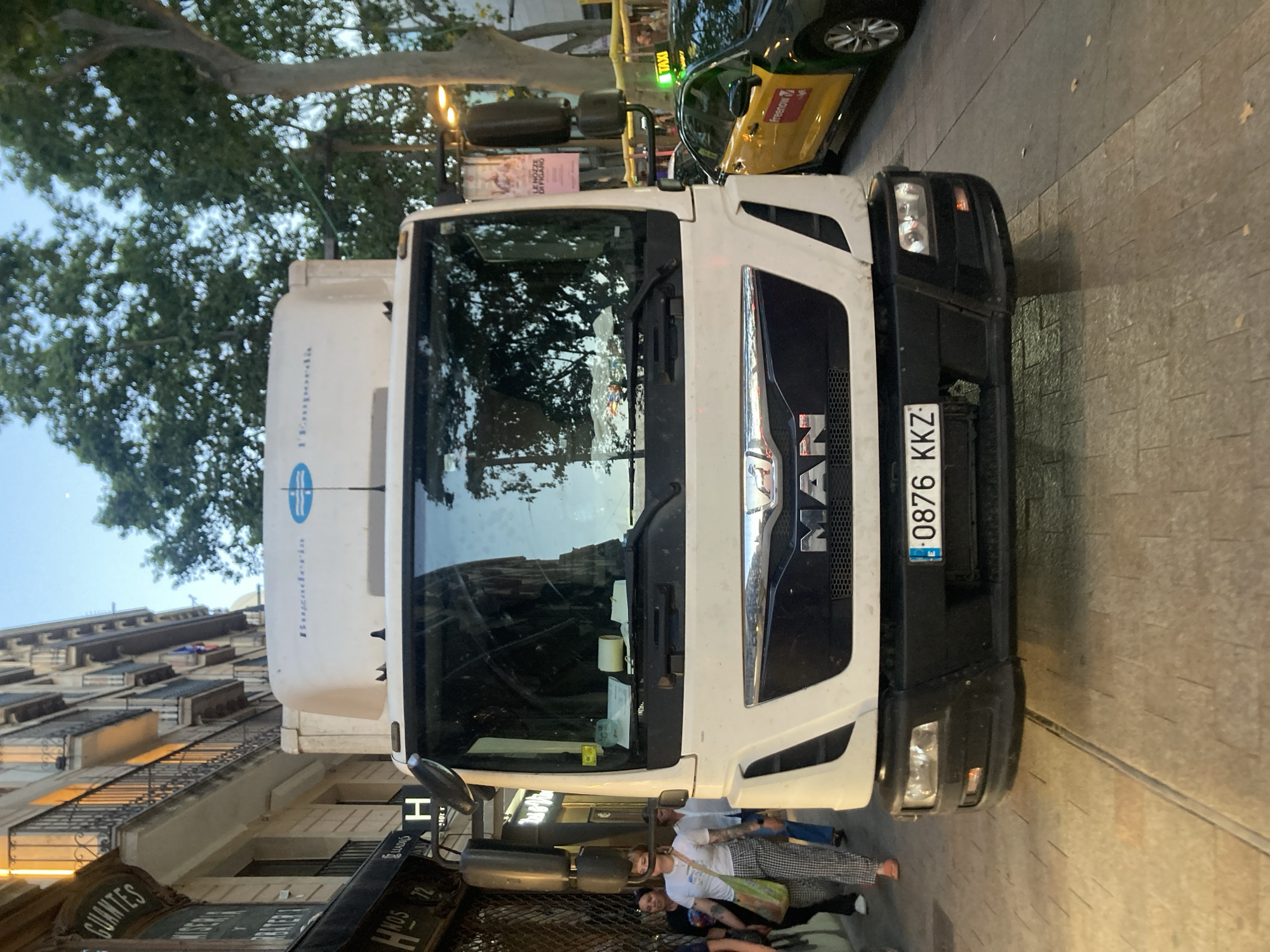 A white MAN truck is parked on a cobblestone street in Barcelona, Spain. The truck has a blue and white logo on its side with the words "Bugaderia" and "l'Empordà". The license plate of the truck reads "0876 KKZ". To the right of the truck, a yellow and black taxi with a "freenow" logo is visible. In the background, there are buildings with balconies and trees. Several pedestrians are walking on the sidewalk in front of the truck. One woman in the foreground is wearing a white t-shirt and checkered pants. The sky is twilight blue.