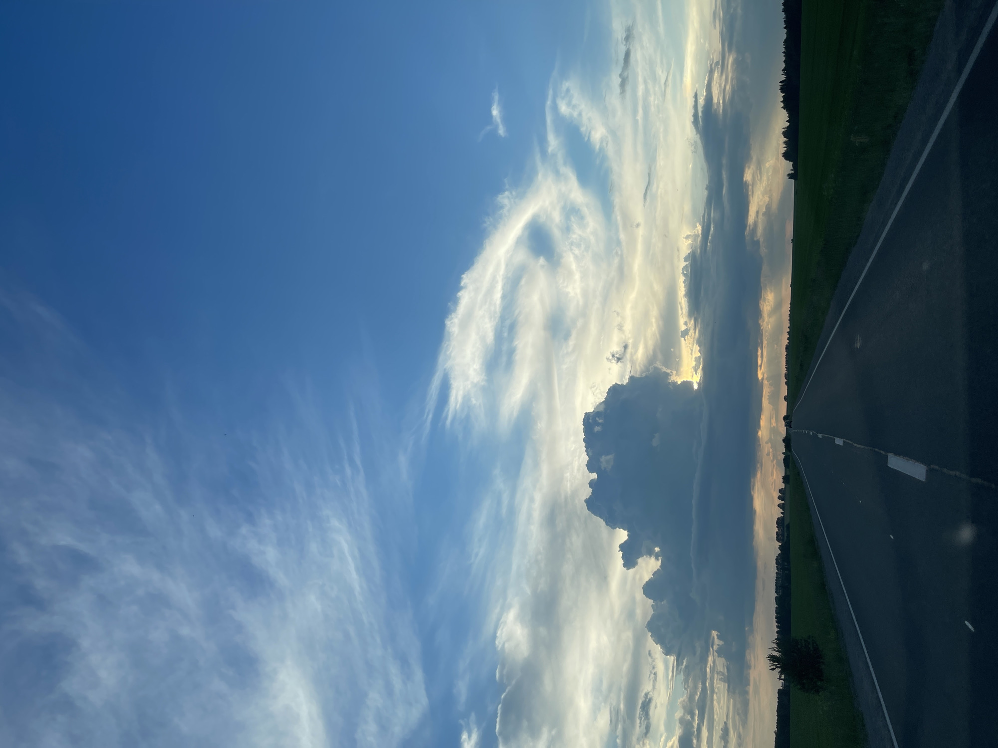An eye-level, medium shot captures a road stretching into the distance, framed by a verdant field on either side. The sky above is a canvas of dramatic clouds, with soft, wispy formations contrasting with a large, dark cumulus cloud dominating the center. The sun peeks through the clouds, casting a warm glow on the horizon. The road, with its white dashed lines, appears empty, with only faint tire marks visible on the asphalt. The surrounding landscape is a tranquil expanse of green fields, with a line of trees forming a dark silhouette against the distant horizon. The overall atmosphere is serene and expansive, evoking a sense of open road and endless possibilities.