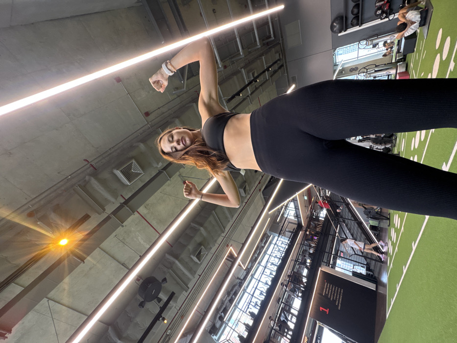 The image displays the interior of a multi-level fitness facility, identified as a Virgin Active gym, located within the city of Bangkok, Thailand. A female subject, appearing to be in her twenties or early thirties, stands centrally in the foreground, facing the camera from a low angle. She has long brown hair and is dressed in a black sports bra and black ribbed leggings. Her arms are bent at the elbows, with fists clenched and raised, indicating a flexing pose. A white wristband is visible on her right wrist.
The floor beneath the subject is green turf marked with white lines, resembling a sports field. In the midground, a multi-story structure with staircases and glass railings extends upwards, showcasing additional gym levels. Several individuals are visible on these upper levels, engaged in various fitness activities, including operating cardio equipment. A black sign with white text, partially legible as "MEMBER LOUNGE," "FREESTYLE," and "GROUP EXERCISE," is positioned on the left side of this structure. Through large windows in the background, a dense urban skyline with high-rise buildings is apparent. To the right, another individual is seen on the green turf performing floor exercises, with a weight rack partially visible behind them. The ceiling features exposed concrete with prominent linear LED light fixtures and a single bright, starburst-effect light source. The overall environment is brightly lit and modern.