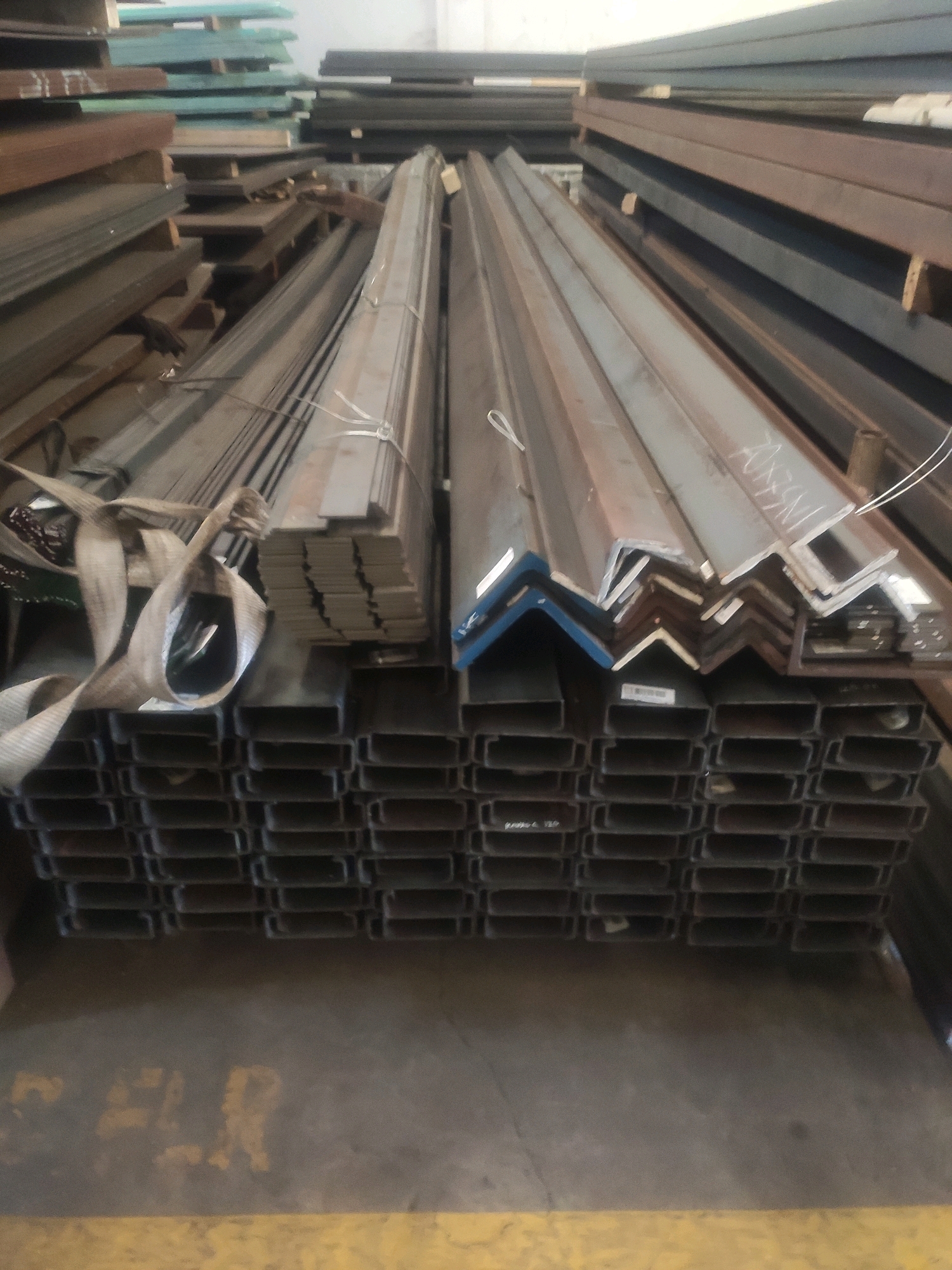An indoor, wide shot shows a storage area with stacks of metal building materials. In the foreground, a large quantity of C-channel steel is neatly stacked in rows. Above these, various lengths of angle iron are stacked in vertical columns. In the background, additional stacks of metal beams and what appear to be sheet metal are visible. The materials are a mixture of steel and possibly aluminum. The environment appears to be a warehouse or industrial storage facility. There are no people visible in the image.