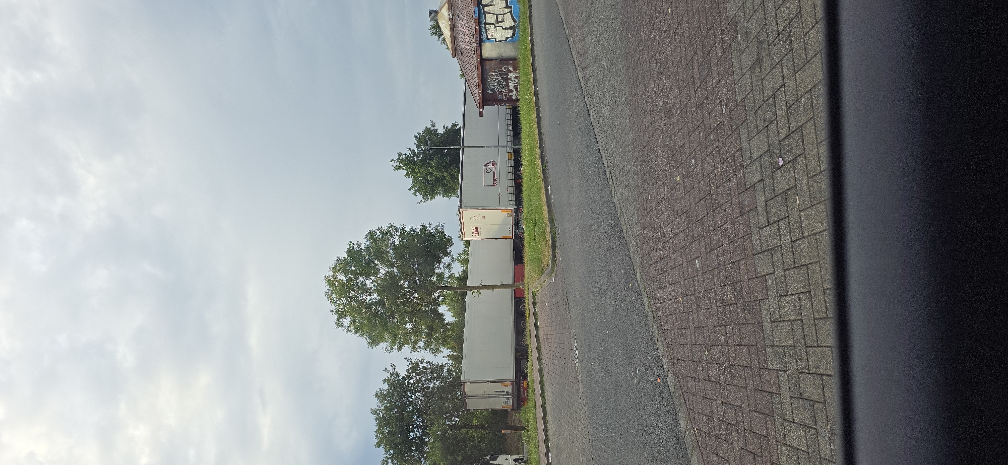 A row of parked semi-trailer trucks is visible on the left side of the image, facing away from the viewer. The trucks are white and appear to be identical in model. Some have logos or writing on their sides, but they are not clearly legible. The trucks are parked on a paved surface next to a grassy area. Behind the trucks, trees and a building with graffiti are visible. The building appears to be an industrial or warehouse structure. The sky is mostly clear with some light clouds. The image is taken from a low angle, possibly from a vehicle. The time of day appears to be daytime, and the weather seems to be mild. The scene is set in an industrial or commercial area, possibly a parking lot or loading dock. There are no people visible in the image. The image appears to be taken in Staufenberg, Germany.