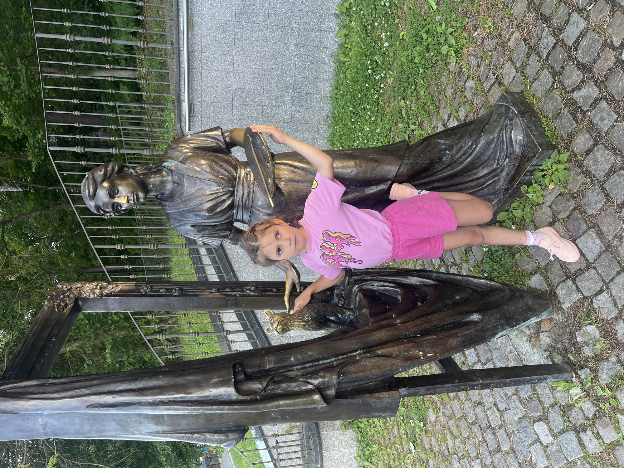 In Svetlogorsk, Russia, a young girl in a pink t-shirt and shorts stands next to a bronze statue of a woman holding a palette and brush. The statue is positioned near a stone walkway and lush greenery. The girl is leaning against the statue, with her right leg bent and foot resting on the base. Her left hand is placed on the statue's dress, and her right hand is extended upwards, mirroring the statue's pose. The statue itself depicts a woman in a classical-style dress, with intricate details in her hair and clothing. Behind the statue, a metal railing borders a grassy area and trees. The overall scene is set outdoors, likely in a park or public square.