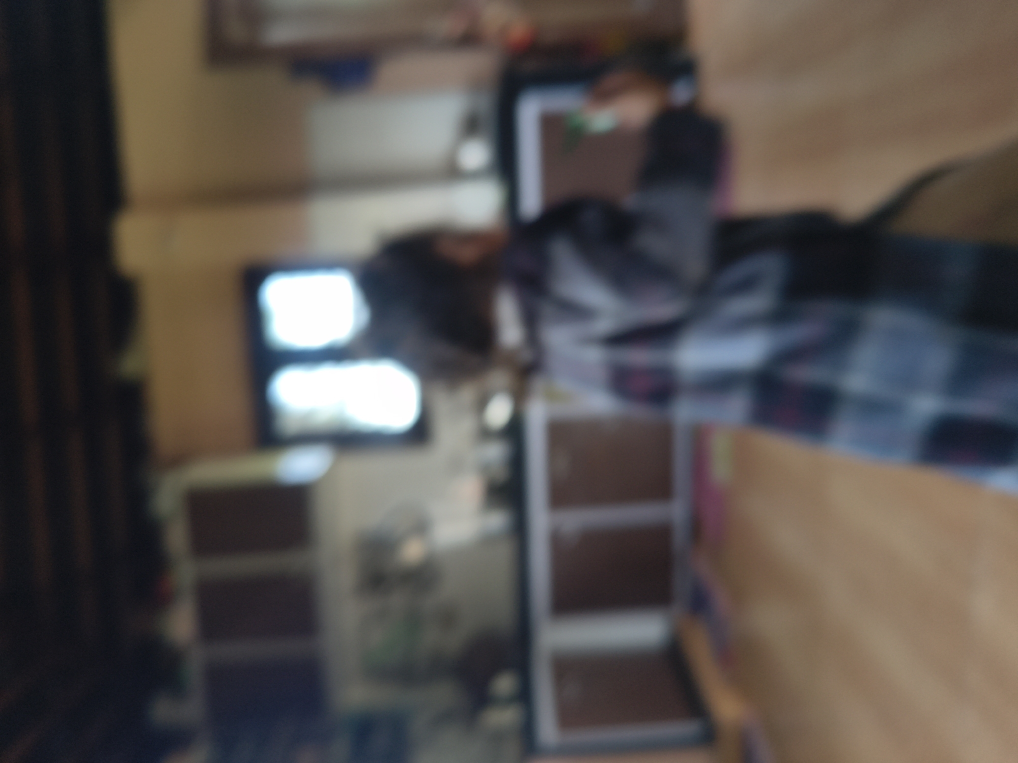 A person is seen from behind, wearing a plaid jacket, in a room that appears to be a kitchen or dining area. The person is holding a rectangular object, possibly a tablet or phone, in their right hand. The room has wooden flooring and cabinets along the wall. The image is blurry, making it difficult to discern finer details. The overall impression is that of an indoor residential setting. The location in the city of Kandi, India is not ascertainable from the visual information present.