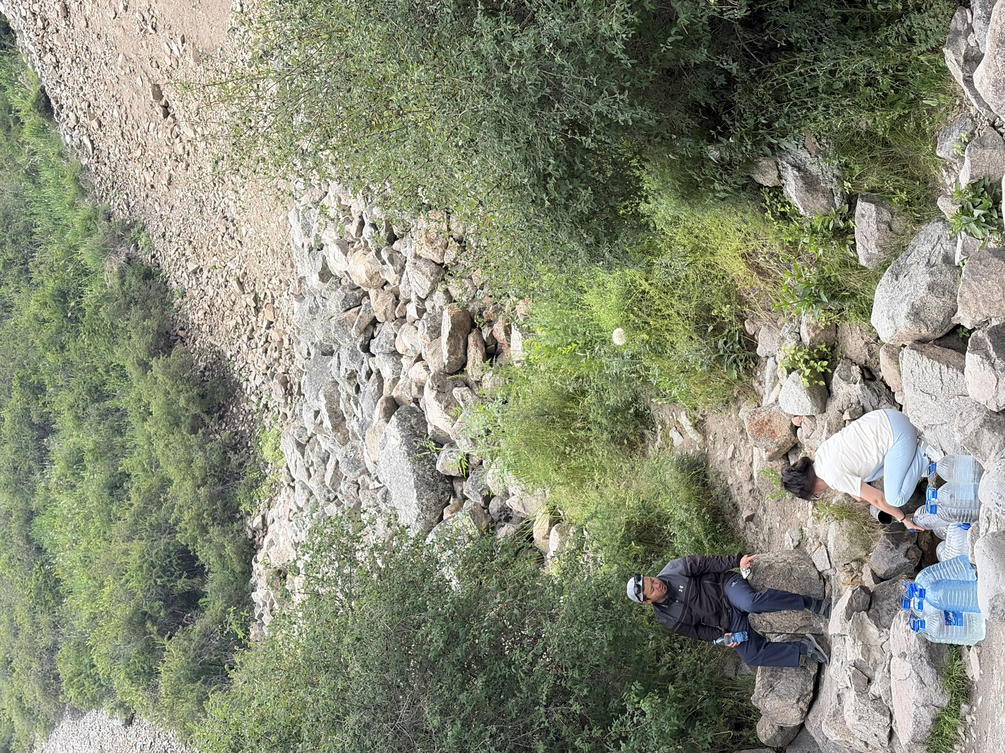 In a natural environment with rocky terrain and abundant green foliage, a man and a woman are actively engaged in filling large plastic water bottles from a natural water source. The man, seated on a rock, holds a bottle of water, while the woman, in a crouching position, directs water from a pipe into several bottles arranged on the ground. The scene suggests a process of collecting fresh water from a natural source, possibly for consumption or transport. The surrounding environment appears to be a wilderness area, with large rocks and dense vegetation dominating the landscape.