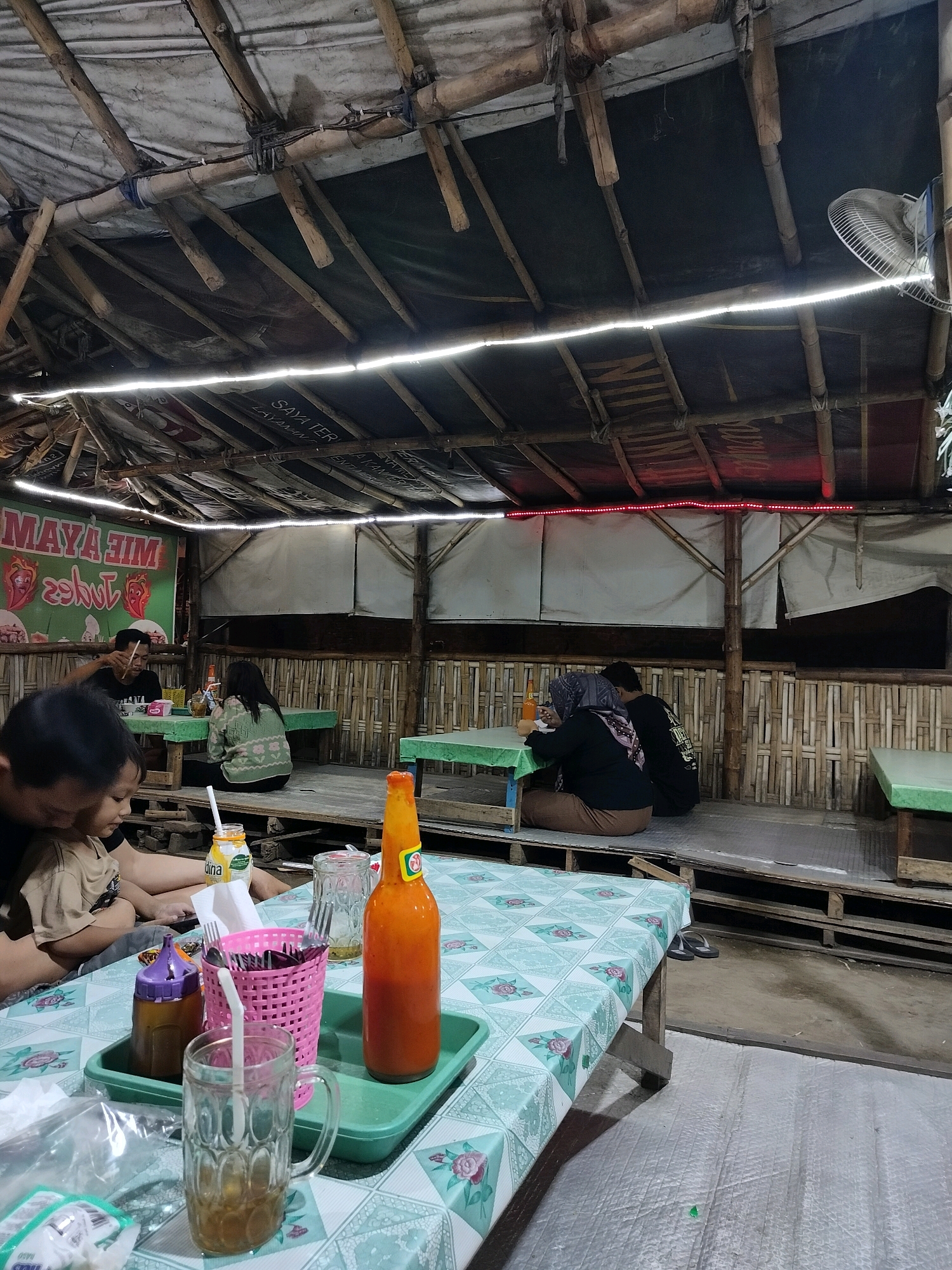 In Trowulan, Indonesia, a casual open-air food stall features a rustic bamboo and tarp roof with string lights illuminating the dining area. Several tables are set up on wooden platforms. In the foreground, a table is covered with a light green tablecloth featuring a floral pattern. On this table, a green plastic tray holds various items including a glass of iced tea with a straw, a pink basket with cutlery, a bottle of orange sauce, and a clear glass with a yellowish liquid. Behind this table, two people, a man and a child, are seated. Further back, another table is occupied by a couple eating. To the right, a bamboo-paneled wall forms the enclosure of the stall. The overall atmosphere is relaxed and informal, typical of local eateries.