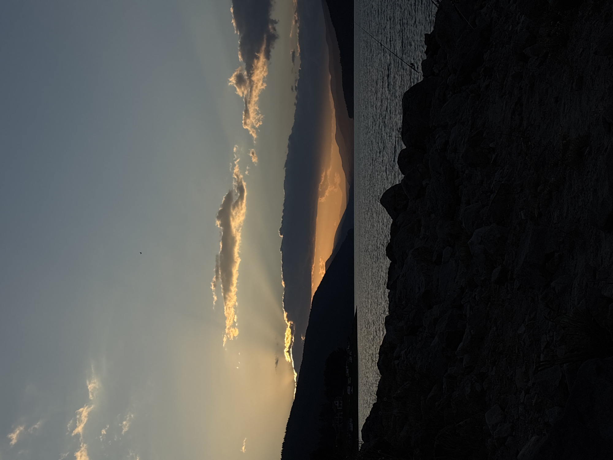 An image taken at dusk captures a serene natural landscape. The foreground is dominated by dark, rocky terrain, with scattered dry grasses. Beyond this, a body of water reflects the dimming light of the sky. In the background, silhouetted mountains rise, their slopes dotted with trees on the left side. Several layers of mountains recede into the distance, fading into a hazy horizon. The sky above is a gradient of pale blue and orange, with scattered clouds illuminated by the setting sun. Sunbeams pierce through a band of clouds, creating a dramatic effect. A single bird is visible in the sky. Two fishing rods are set up on the rocky shore on the right side of the image.