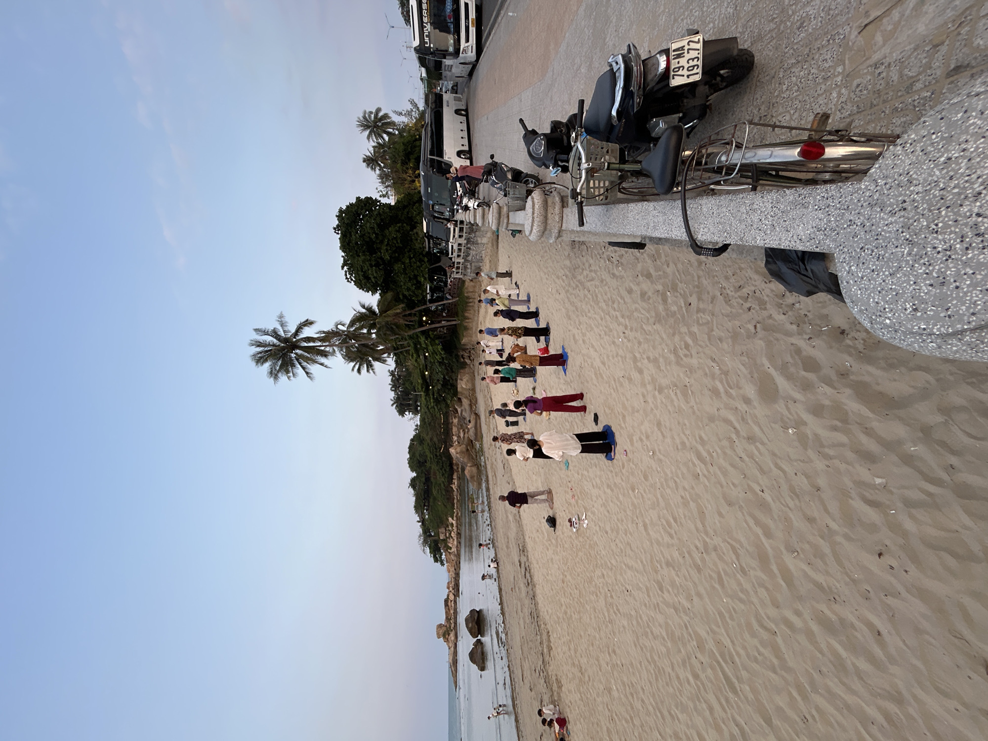 The image depicts an early morning or late afternoon scene on a beach in Bắc Nha Trang, Vietnam. The setting includes a sandy beach, calm ocean water with visible rocks, and a paved road or walkway separated from the beach by a low concrete barrier. Lush green vegetation and several palm trees line the area above the beach.
A group of approximately 15-20 people are engaged in an organized activity on the sandy beach, likely a yoga or exercise class. Many participants are positioned on colorful mats (blue, green, red, purple) or standing in various poses, generally facing towards the water. A few other individuals are visible closer to the shoreline.
Along the paved road, several vehicles are parked. These include multiple large tour buses, one of which has "UNIVERSE" visible on its side. Numerous motorbikes and scooters are also parked, with one prominent in the foreground. The license plate of this motorbike displays "79-NA 193.72". The sky is a clear, light blue with subtle cloud formations, indicating soft natural lighting. The overall atmosphere appears calm and focused, with individuals participating in their activity.