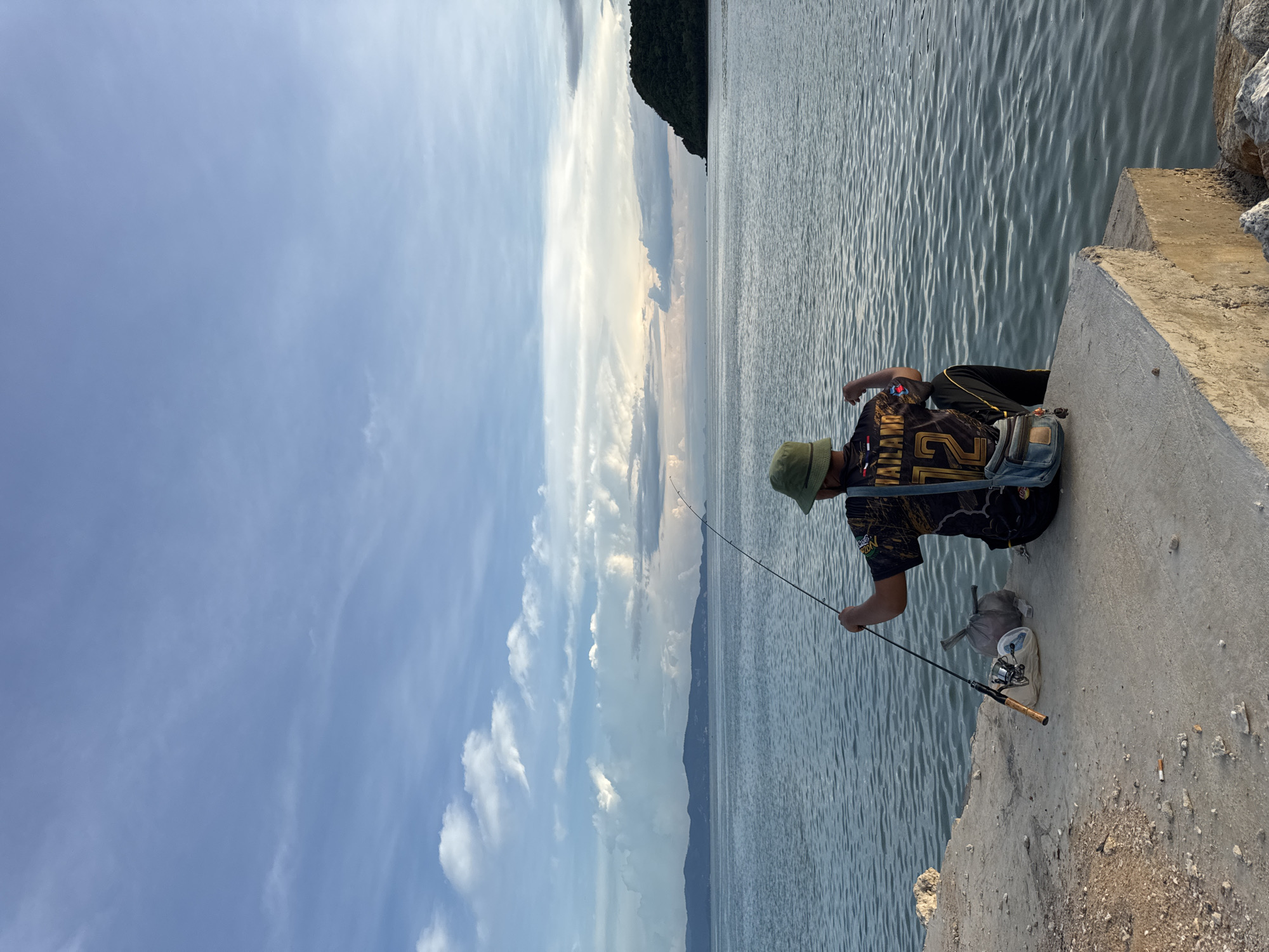 A man is fishing on a concrete pier in Koh Phangan, Thailand. He is wearing a black t-shirt with "THAILAND 12" in yellow lettering and a green bucket hat. He is holding a fishing rod and appears to be looking out at the sea. Next to him are a fishing reel, a beige bag, and a blue fanny pack. The sea is calm with gentle waves. In the distance, there are islands with green vegetation and a cloudy sky with hues of pink and orange, suggesting it is either sunrise or sunset. The concrete pier is rough and shows signs of wear.