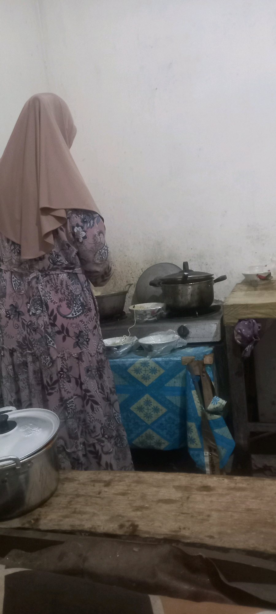 In a dimly lit indoor setting, likely a kitchen in Pontang, Indonesia, a person wearing a light brown hijab and a patterned dress is standing. The person is facing away from the camera, towards a stovetop where a pot with a lid is placed. The stovetop is a small, freestanding unit with a metal surface. To the right of the stovetop, a wooden shelf holds a bowl and a red object, possibly food. A blue and yellow patterned cloth is draped over a surface in front of the stovetop, with some utensils or bags attached to it. In the foreground, a large metal pot with a lid is visible on a wooden surface. The walls appear to be rough and stained, suggesting a rustic or utilitarian environment.