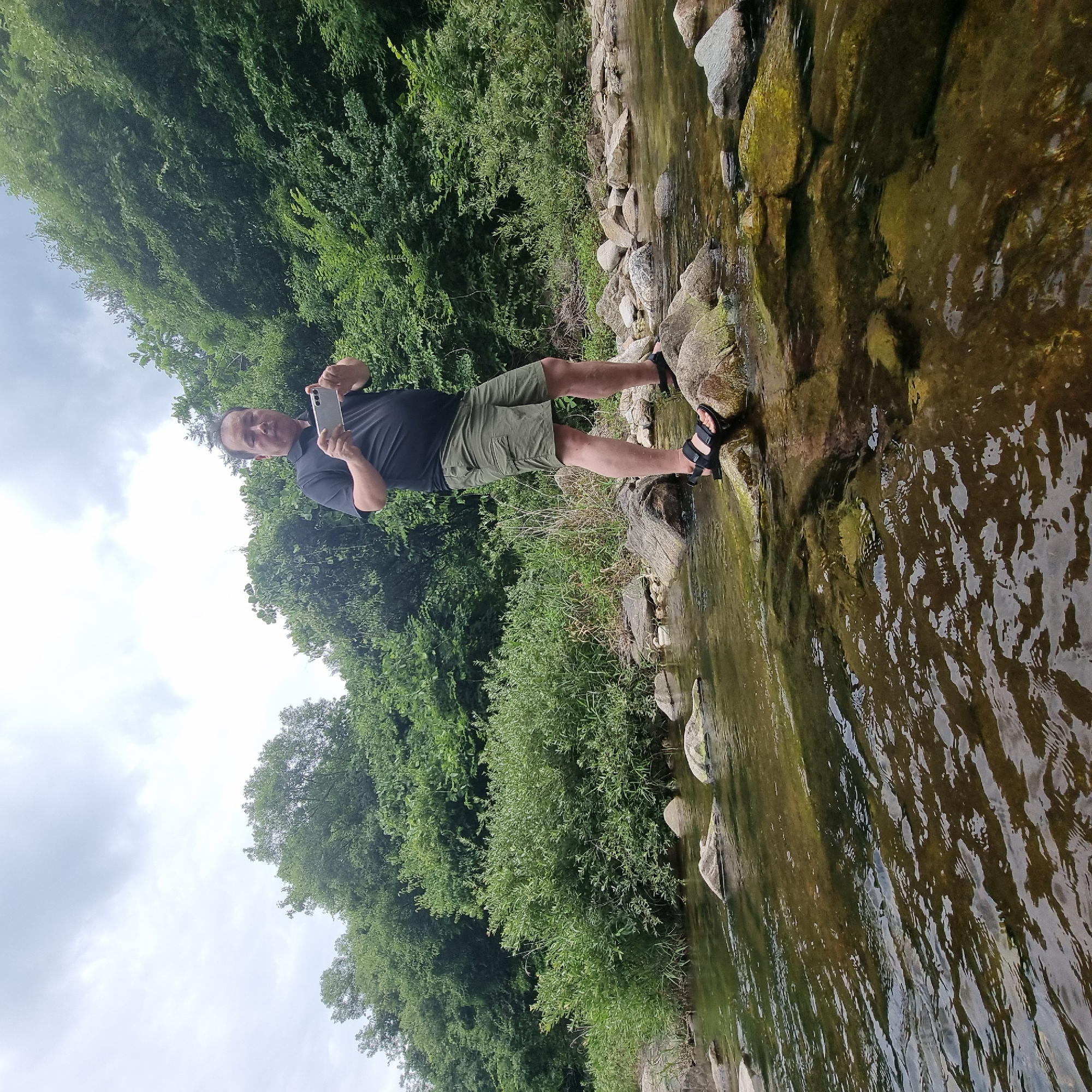 A man is standing on rocks in a shallow, clear river. He is wearing a black polo shirt and khaki shorts, and black sandals. He is holding a smartphone in his hands, as if taking a picture or recording a video. The riverbed is visible through the water and is covered with rocks and pebbles. Lush green trees and bushes line the banks of the river, creating a natural, serene environment. The sky is overcast with gray clouds. The scene appears to be in a natural outdoor setting, possibly a park or a hiking trail. The location of Mureungdowon-myeon, South Korea, suggests a rural or mountainous area with natural beauty, consistent with the image.