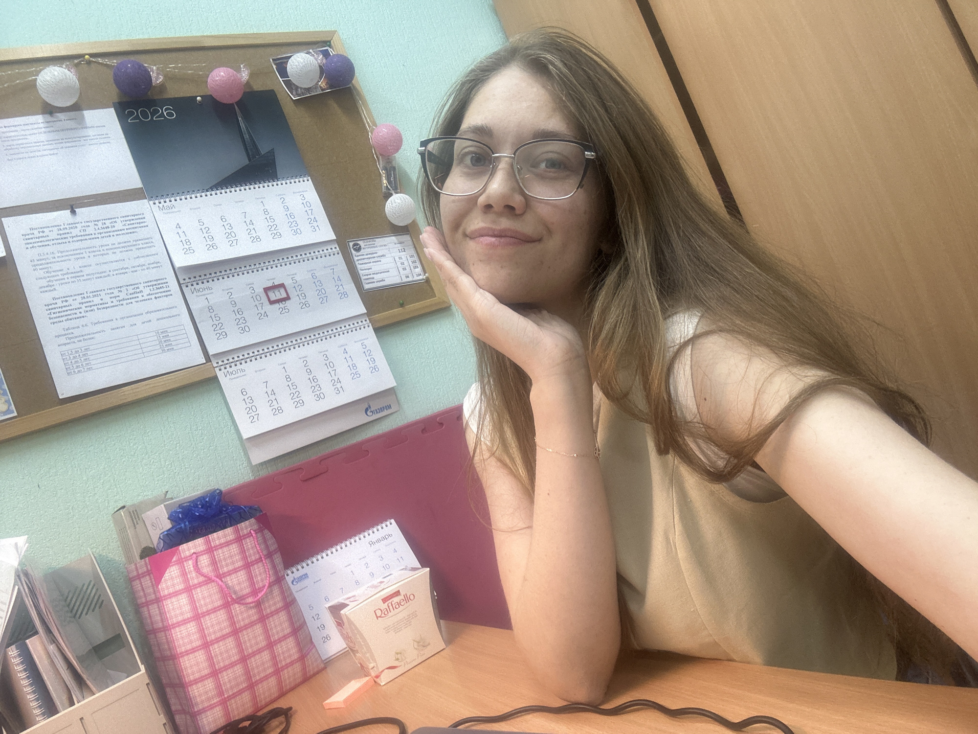 A young woman with long brown hair and glasses is sitting at a wooden desk. She is looking at the camera and resting her chin on her hand. She is wearing a light-colored sleeveless top. Behind her, a bulletin board is covered with various documents, including calendars for 2026, and decorative balls. To her left, a pink plaid gift bag and a box of chocolates are visible on the desk. A red mat is behind the gift bag, and a wire is visible in the foreground of the desk. The overall environment appears to be an office or study space. The presence of calendars with Russian text and the visible brand names on some documents suggest the location is likely in Russia, specifically in the city of Chaikovsky, Russia.