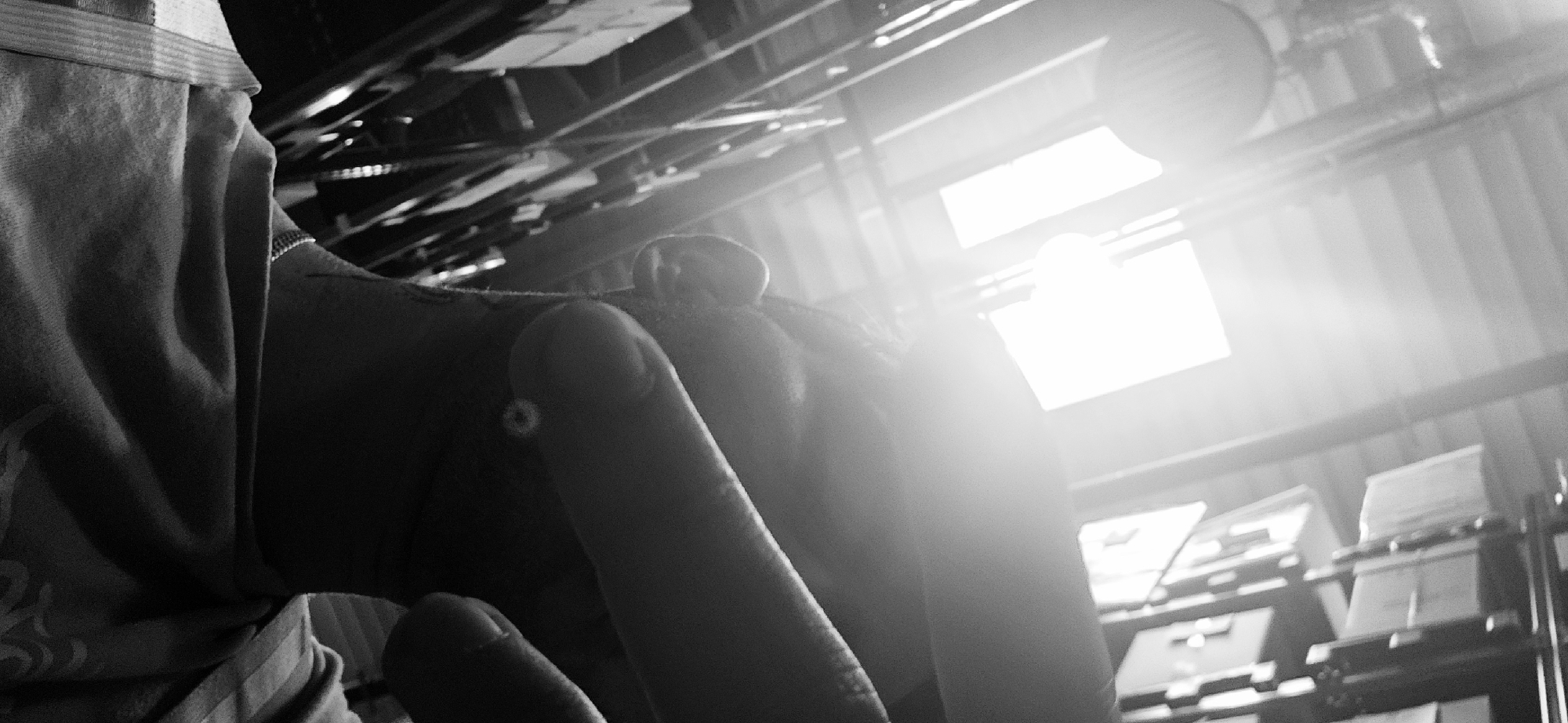 The monochrome image, shot from a low angle, depicts a person's hand in the foreground, obscuring part of the view. The hand appears to be holding or reaching towards something, but the exact action is unclear due to the close proximity and limited view. The background is brightly lit by sunlight entering through windows, suggesting an indoor setting. The architecture seems industrial, with beams, pipes, and corrugated metal visible in the ceiling and upper portions of the frame. There is shelving with boxes and papers in the background. No definitive landmarks are recognizable to definitively place the location in Plovdiv, Bulgaria.