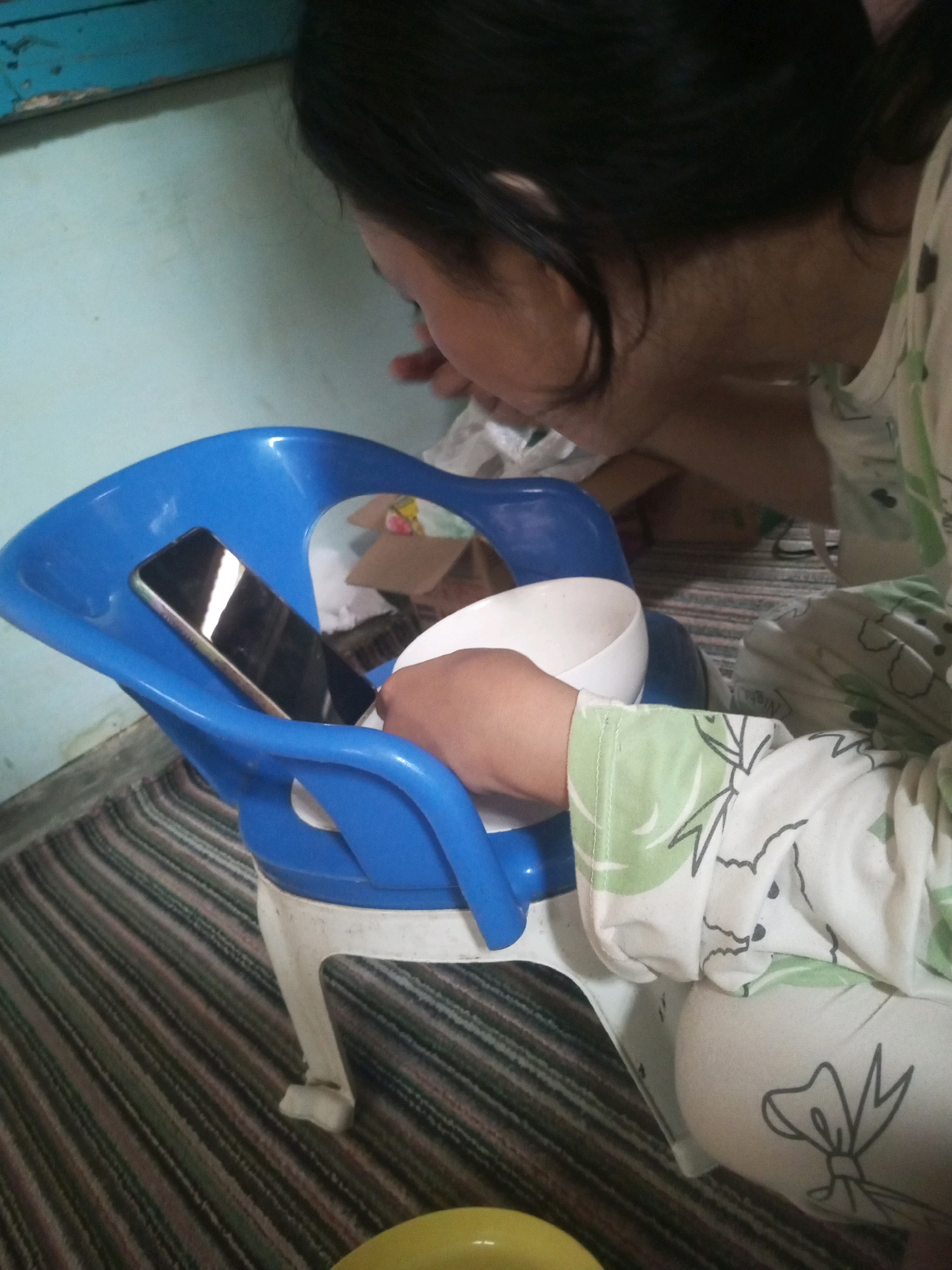 In Andongsili, Indonesia, a close-up shot captures a person's arm resting on a blue plastic chair. A dark-colored smartphone lies on the seat of the chair, reflecting light. A white bowl is positioned next to the arm, partially obscuring the chair's interior. The person wears a garment with a green and white floral pattern. The background shows a striped carpet and a teal-colored wall.