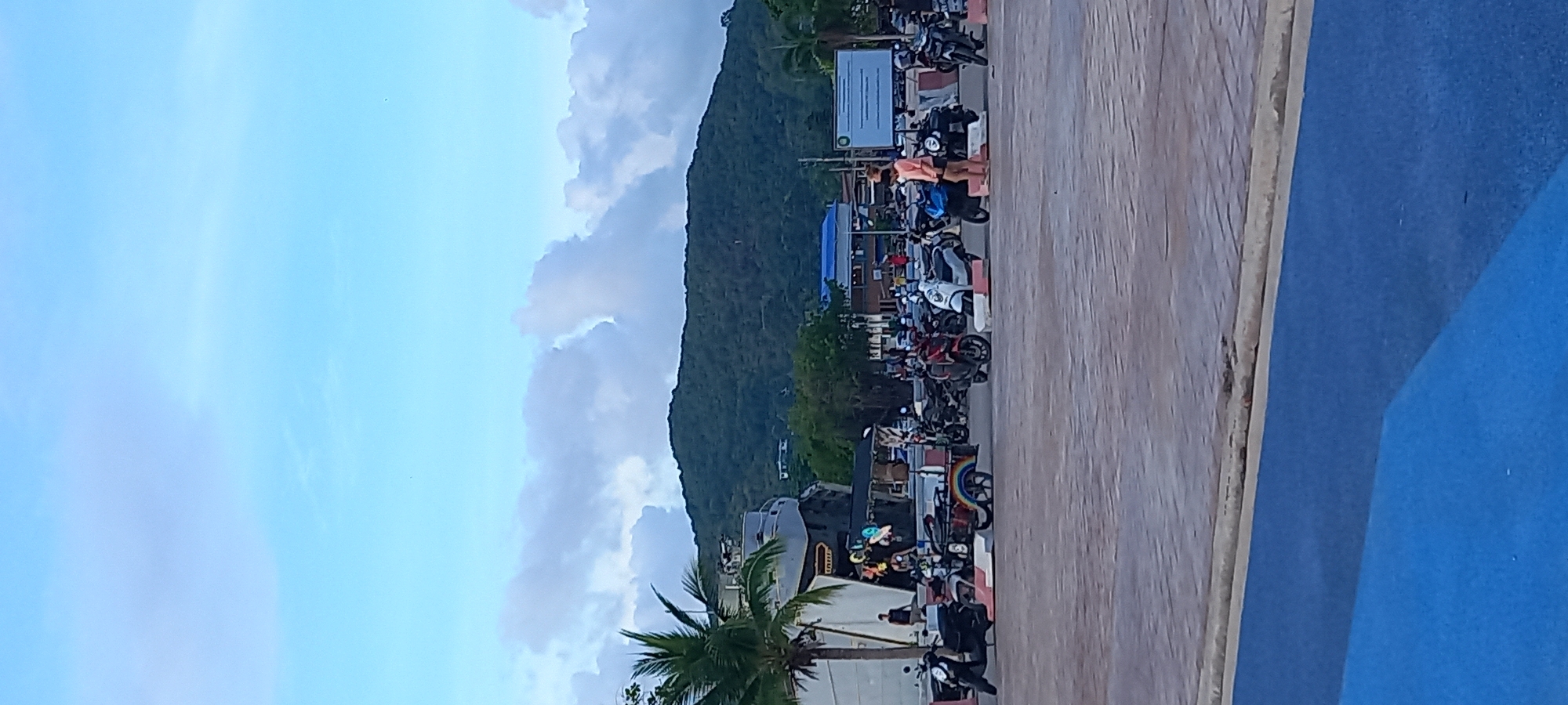 The image depicts a scene in Thong Sala, Thailand, featuring a street lined with parked motorbikes and a wooded hillside. The sky above is partly cloudy. A wooden walkway or boardwalk runs along the right side of the image, adjacent to a blue surface, possibly a road or painted area. On the left of the parked motorbikes, there is a mix of buildings, including one with a blue facade and a sign with lettering. A palm tree is visible at the bottom left corner. Two people are standing near the center of the row of parked motorbikes.