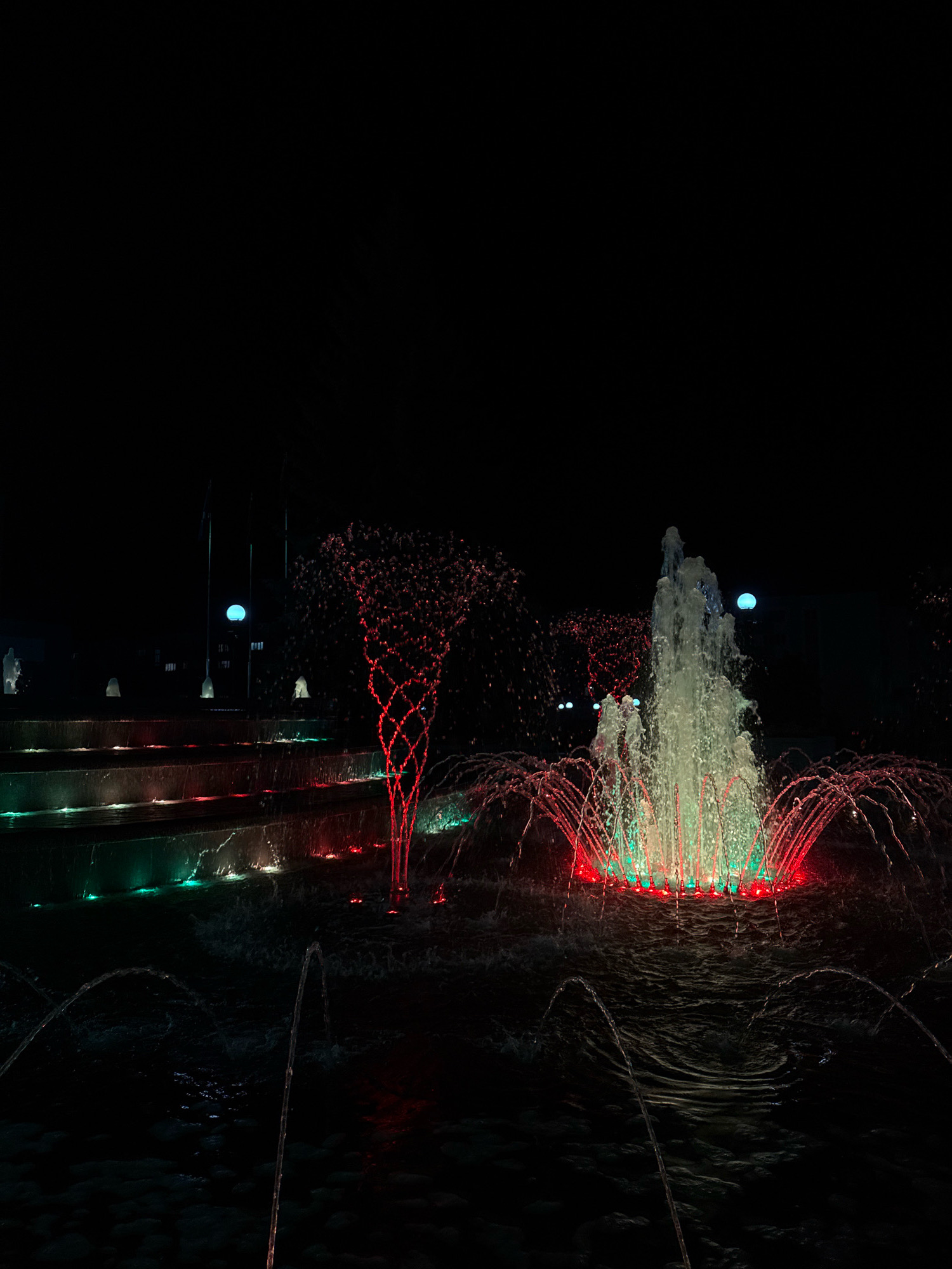 The image depicts a nighttime scene of an illuminated fountain in Mikashevichy, Belarus. The fountain features multiple tiers with water cascading down. The water is lit with red and green lights, creating a vibrant display against the dark background. In the background, there are buildings and flagpoles, suggesting an urban or public space. The fountain is the central subject, with water jets creating a dynamic and artistic effect. There are no people visible in the image. The overall atmosphere is serene and visually striking due to the interplay of light and water.