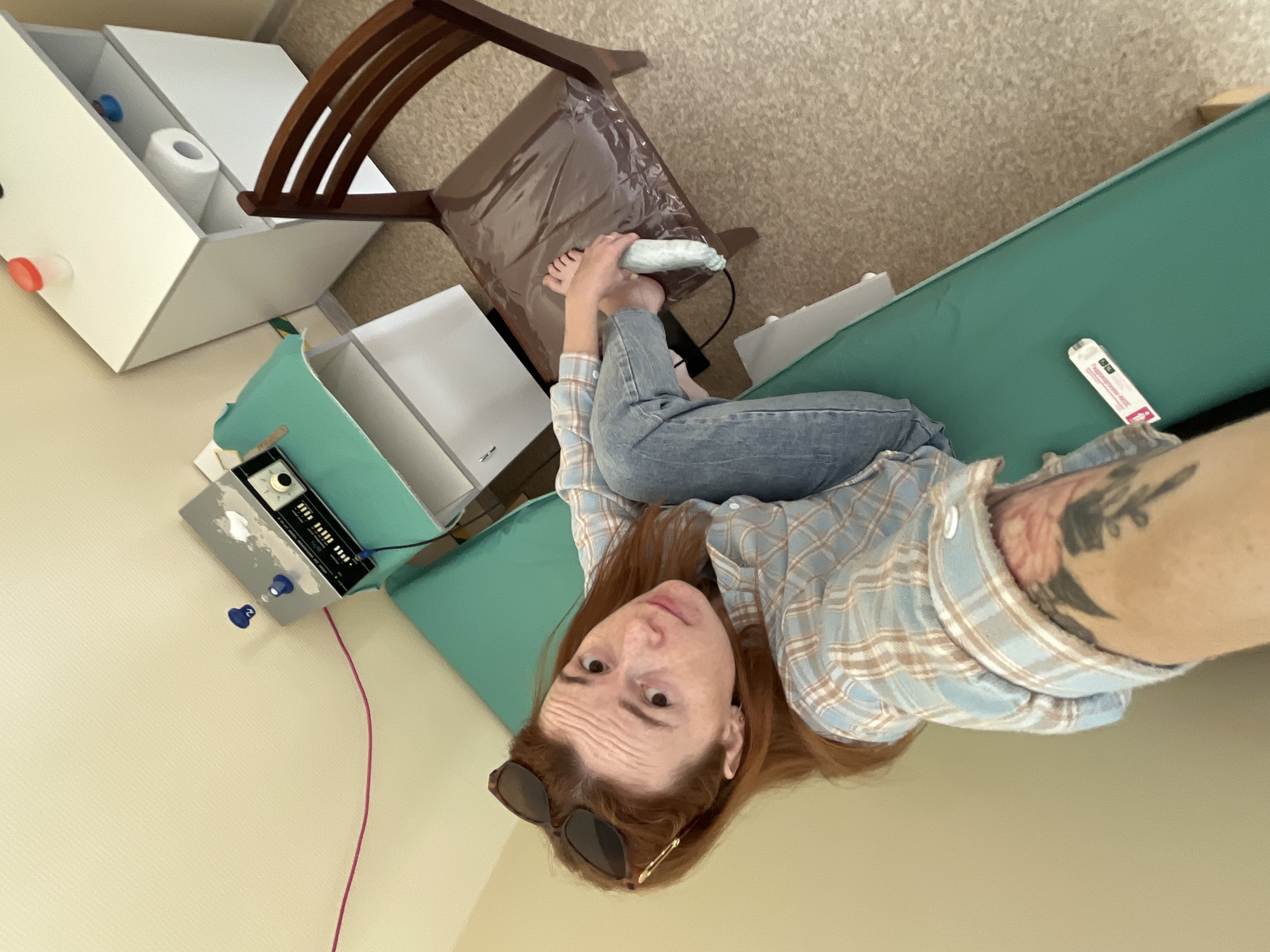 A young woman with long reddish-brown hair is seen lying on her back on a green examination table, looking directly at the camera. She wears a light blue and beige plaid shirt, blue jeans, and sunglasses are pushed up onto her forehead. A tattoo with dark and reddish elements is visible on her right upper arm. Her expression appears neutral, with slightly raised eyebrows.
Her bare feet are visible, resting on a brown chair with a plastic covering, and she is holding a white, soft-looking object between her feet. To her left, on a lower surface, is a black and silver device with several dials and a label that reads "BEEH TRONIC," connected by a dark wire and a pink wire.
On the green table next to her, a white packet or tube is visible with the text "Pregnancy-test" and "Определяет беременно" (Determines pregnancy) printed on it. In the background, to the left, are white shelving units containing items like a roll of toilet paper and small containers. A wooden chair is also present near the shelving. The floor appears to be carpeted. The overall setting suggests an indoor environment, possibly a medical or therapeutic clinic.
