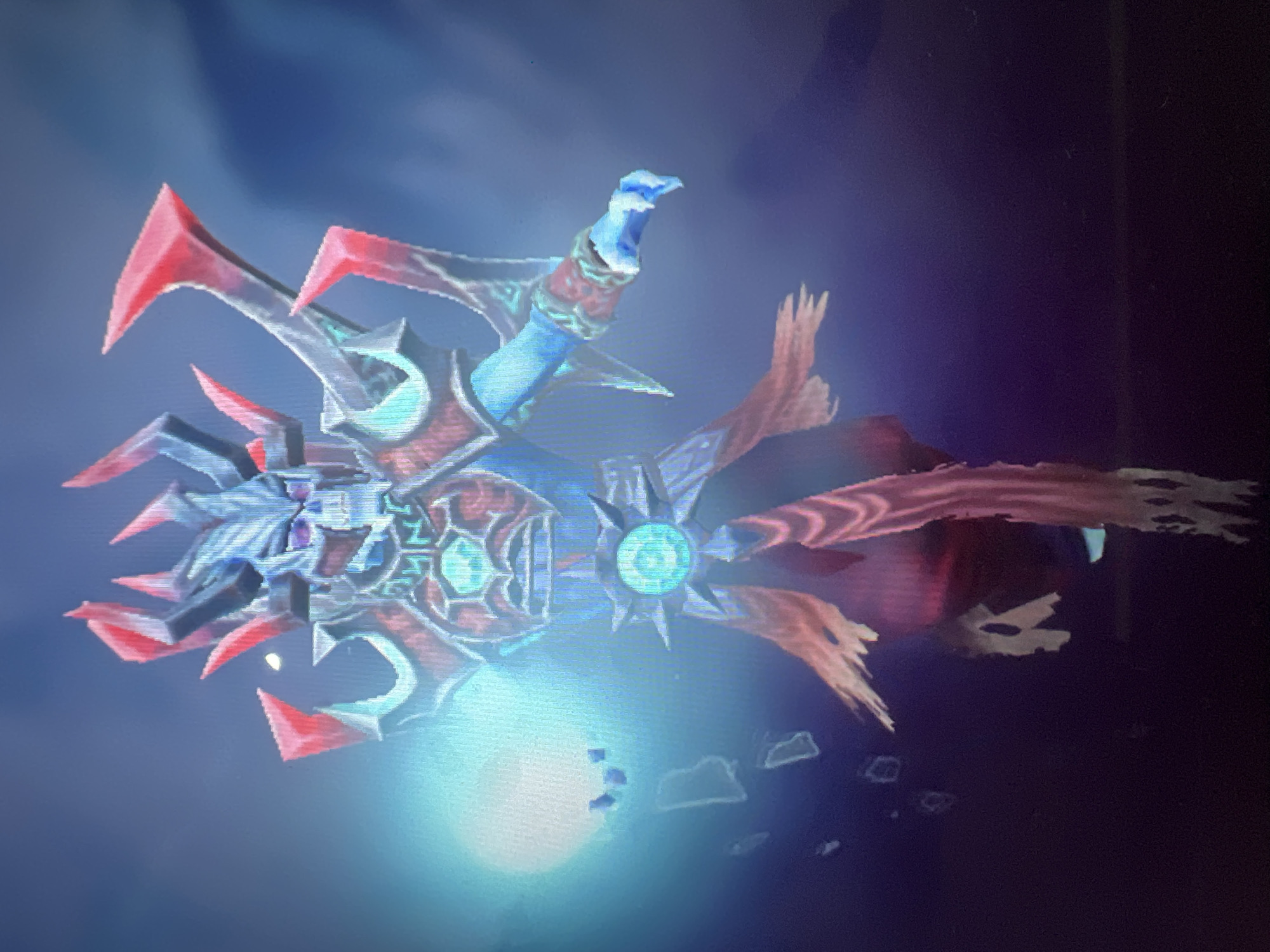 A digital rendering depicts a skeletal, blue-skinned humanoid figure adorned with ornate armor. The figure's head is a skull with large, pointed horns, some of which are tipped in red. Its chest plate features a glowing, teal gem surrounded by intricate carvings. The figure's left hand emits a bright, white light, and a series of small, floating, irregular shapes are visible around it. The figure's lower body transitions into flowing, tattered robes or tendrils of a reddish-pink hue, with some parts appearing frayed and ghostly white. The background is a dark, blurred gradient of blue and purple, suggesting a mystical or otherworldly setting. The image appears to be a screenshot from a video game or digital artwork. There is no discernible connection to the city of Đà Nẵng, Vietnam.