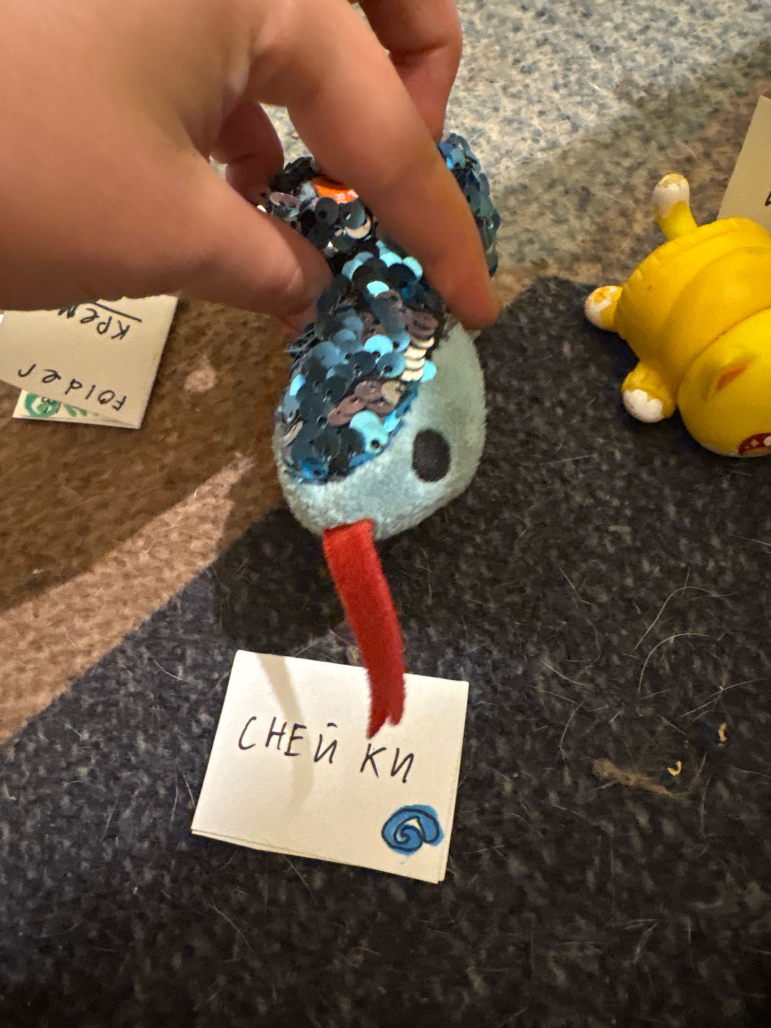A close-up, overhead shot shows a hand holding a small, plush toy with blue and silver sequins. The toy has a light blue body, a black eye, and a red, pipe-cleaner-like appendage extending from its mouth. A small, white piece of paper with handwritten Cyrillic text and a blue swirl drawing is placed in front of the toy. To the right of the toy, a bright yellow, cartoonish plastic figure resembling a cat is lying on its back. The scene is set on a textured, dark grey surface, possibly a rug or carpet. In the background, a portion of a light-colored fabric with a pattern is visible. The overall lighting suggests an indoor setting. There are no identifiable landmarks or indications of the specific city of Ashvem, India in this image.