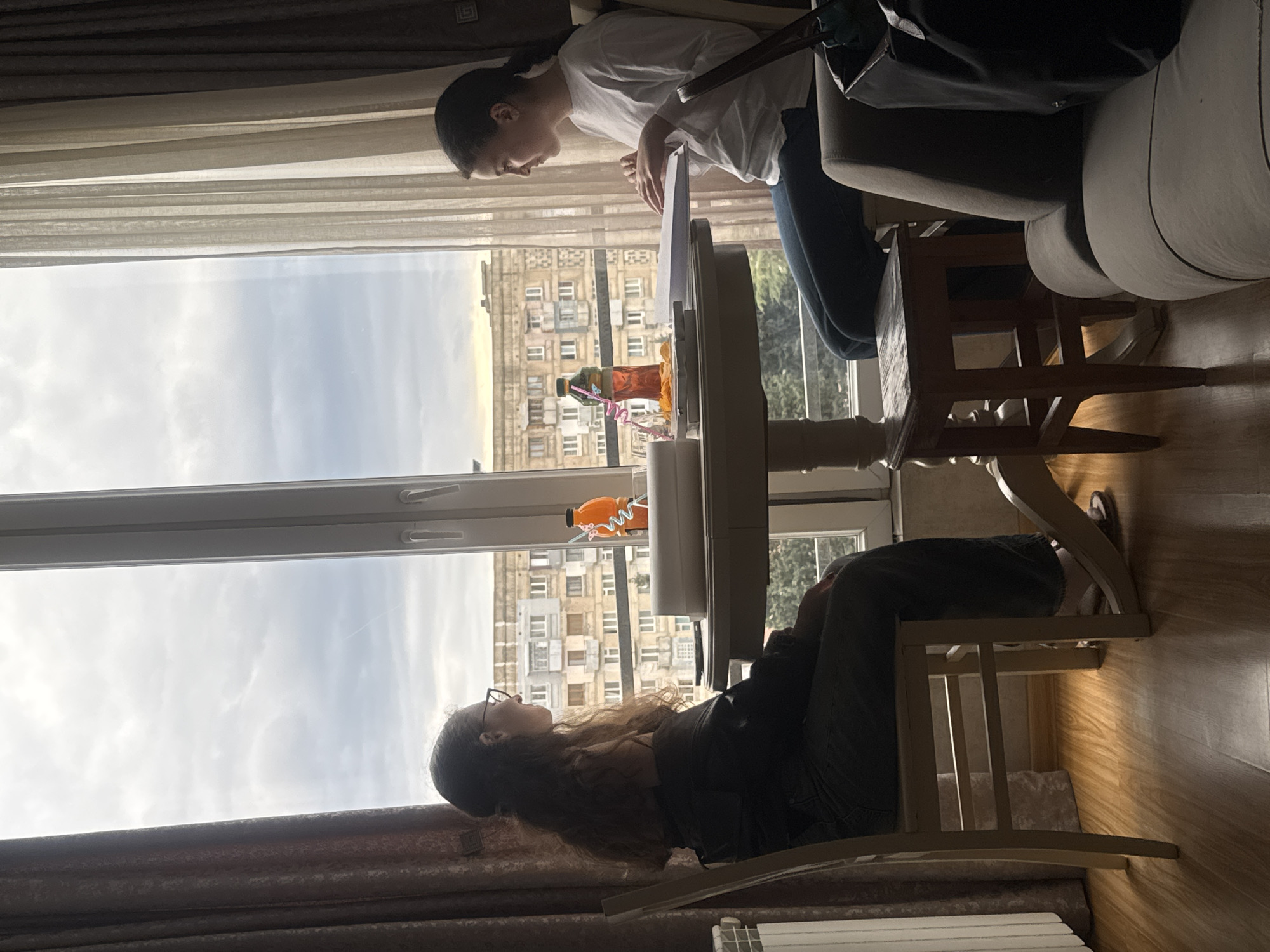 Two people are positioned at a table by a window. One person, wearing a white shirt and jeans, is standing and leaning over the table, while the other, wearing glasses and a dark-colored top, is sitting. The location is likely an apartment with a view of a large building, possibly in Tbilisi, Georgia. A window is in the background, overlooking the urban landscape. A bottle is on the window frame.