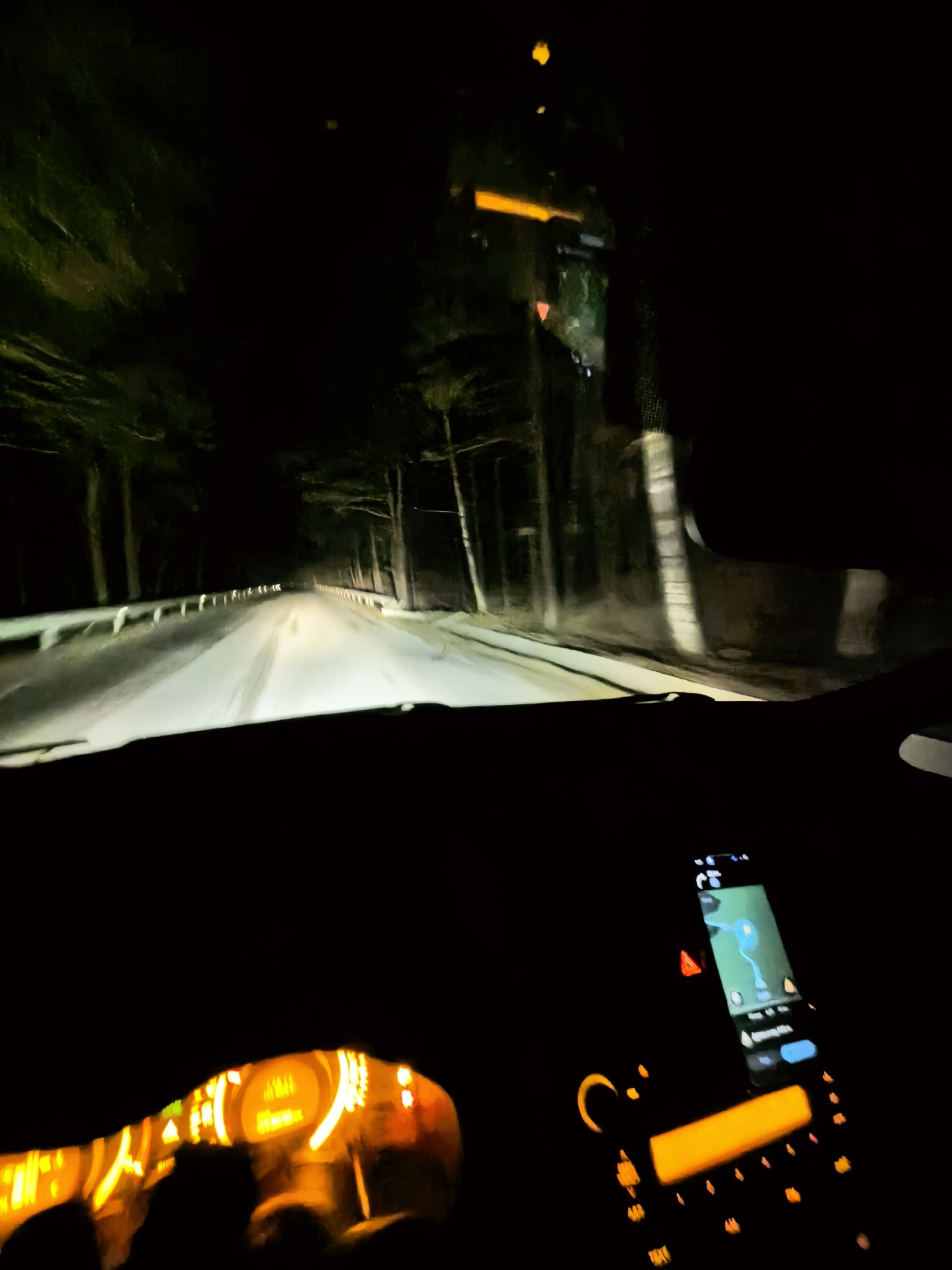 The image depicts a night scene from inside a car driving on a road bordered by trees. The road appears to be illuminated by the car's headlights. The dashboard is visible, with the speedometer and other gauges dimly lit, along with a phone showing a navigation app. The location is Unknown, Unknown. There are no visible people.