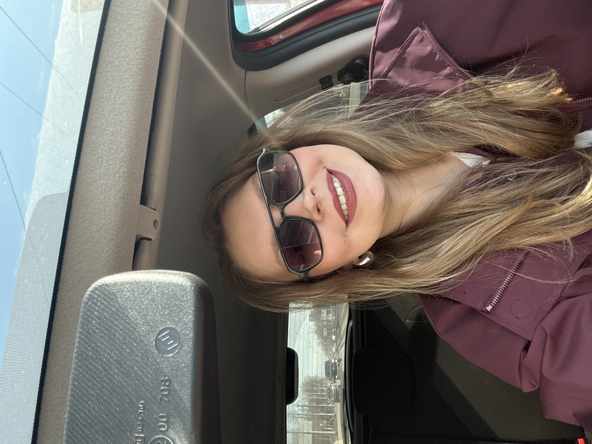 A young woman with long blonde hair, wearing sunglasses and a maroon jacket, is in the driver's seat of a car. The car appears to be moving along a city street. Through the rear-view mirror, other vehicles and a skyline can be seen. The location is inferred to be Izhevsk, Russia.