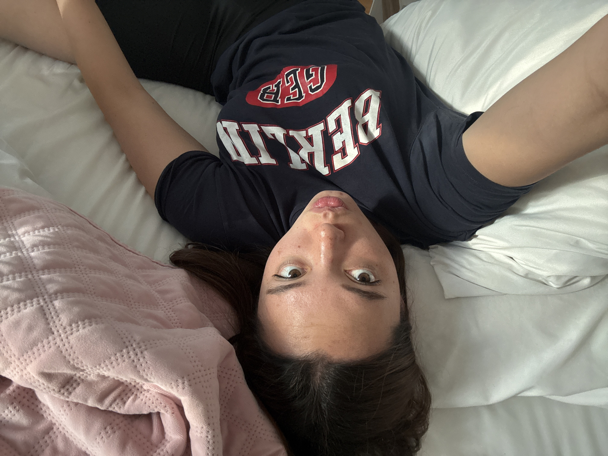 A person is lying on their back on a bed, wearing a dark blue t-shirt with the word "BERLIN" in large white lettering and a red logo above it. The person has brown hair and their eyes are wide open, looking upwards. The background consists of a white pillow and a pink quilt with a quilted pattern. The person is lying on a bed. The location of the scene is indeterminable, as there is no sign to identify San Bernardino Tlaxcalancingo, Mexico, and there are no objects or signs that might indicate it.