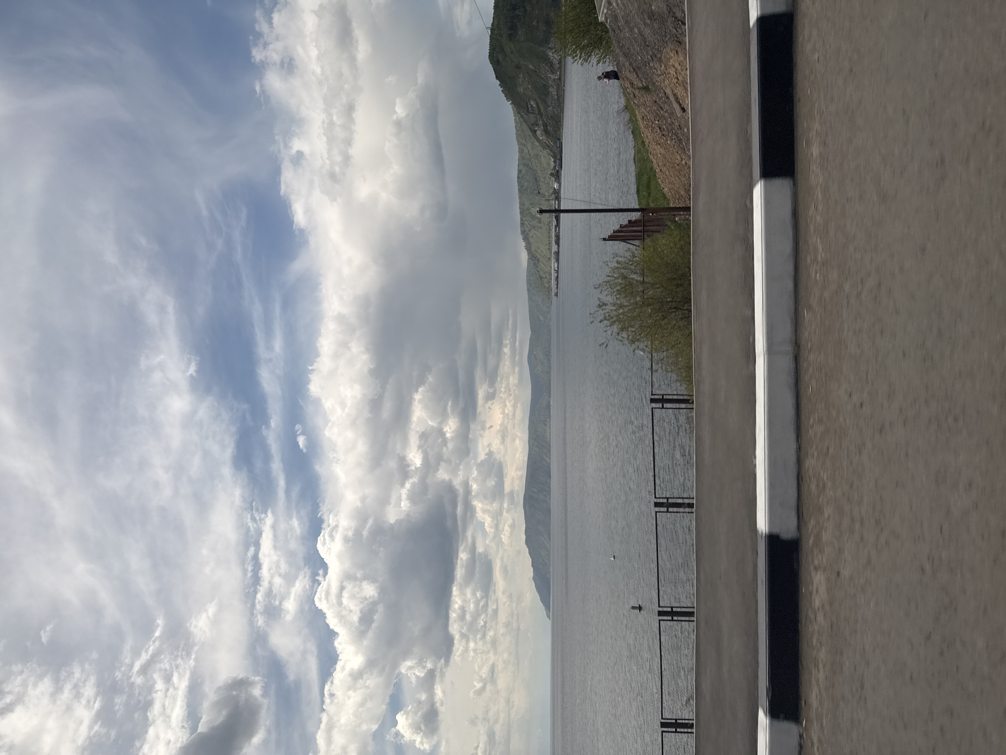 The image captures a cloudy sky and a lake landscape in Listvyanka, Russia. The sky is partially blue, with layers of white and grey clouds. Adjacent to the water is a rocky hillside with vegetation. A black and white barrier is visible on the right side of the image. The lake appears calm, with a few distant structures visible along the shore. An overhead power line is present as well.