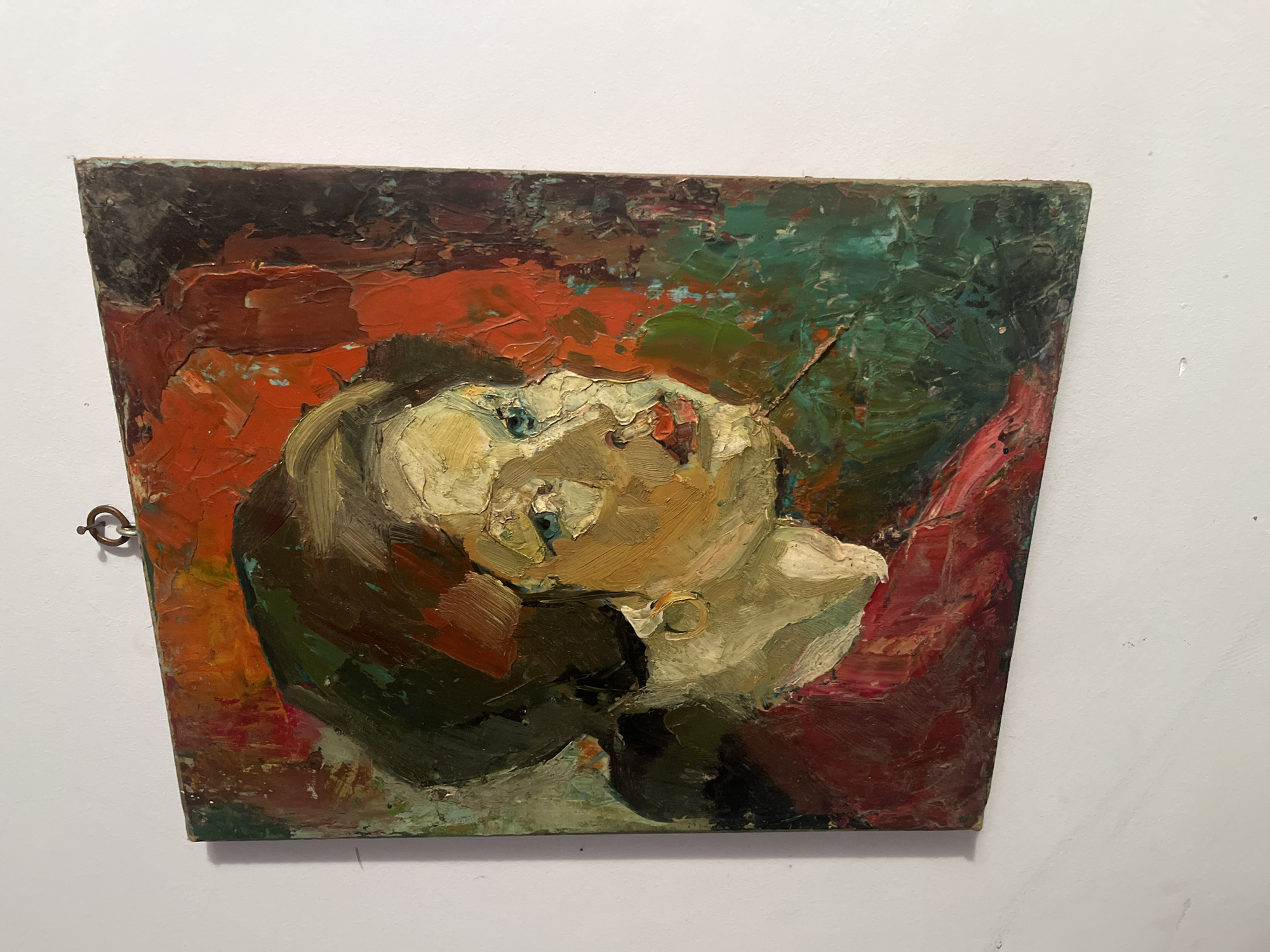 The image depicts a small, rectangular oil painting hanging on a white wall. The painting features a heavily impasto portrait of a woman with pale yellow skin, blue eyes, and red lips. She is facing upwards and slightly to the left. Her hair is dark brown or black, and she appears to be wearing an earring. The background consists of textured brushstrokes in shades of red, orange, green, and dark brown. A metal ring is attached to the left side of the painting, presumably for hanging. The location of the image in Goce Delcev, Bulgaria is not discernible from the visual content.