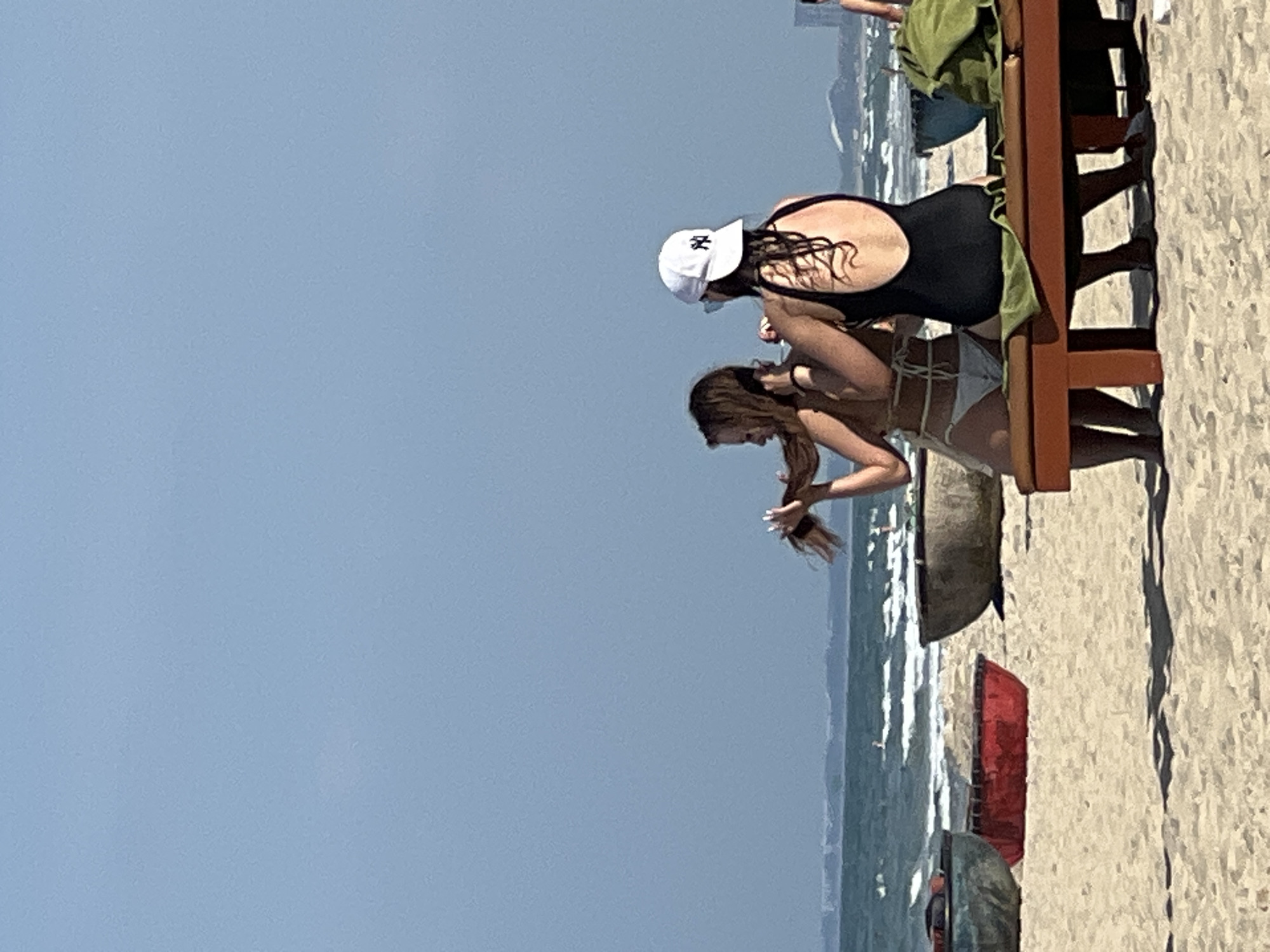 Two women are seen on a beach in Đà Nẵng, Vietnam, sitting on a wooden lounge chair. The woman on the left, facing away from the camera, wears a light-colored bikini and has her hair down. The woman on the right, also facing away, wears a black one-piece swimsuit and a white baseball cap with a black logo. She is seen adjusting the hair of the woman on the left. In the background, the ocean with gentle waves is visible, and further out, the coastline of Đà Nẵng city can be seen. Several round, traditional Vietnamese fishing boats, known as "thúng chài" or basket boats, are resting on the sand in the middle ground, some with colorful interiors. The sand is light-colored and appears dry. The sky is clear and a light blue.