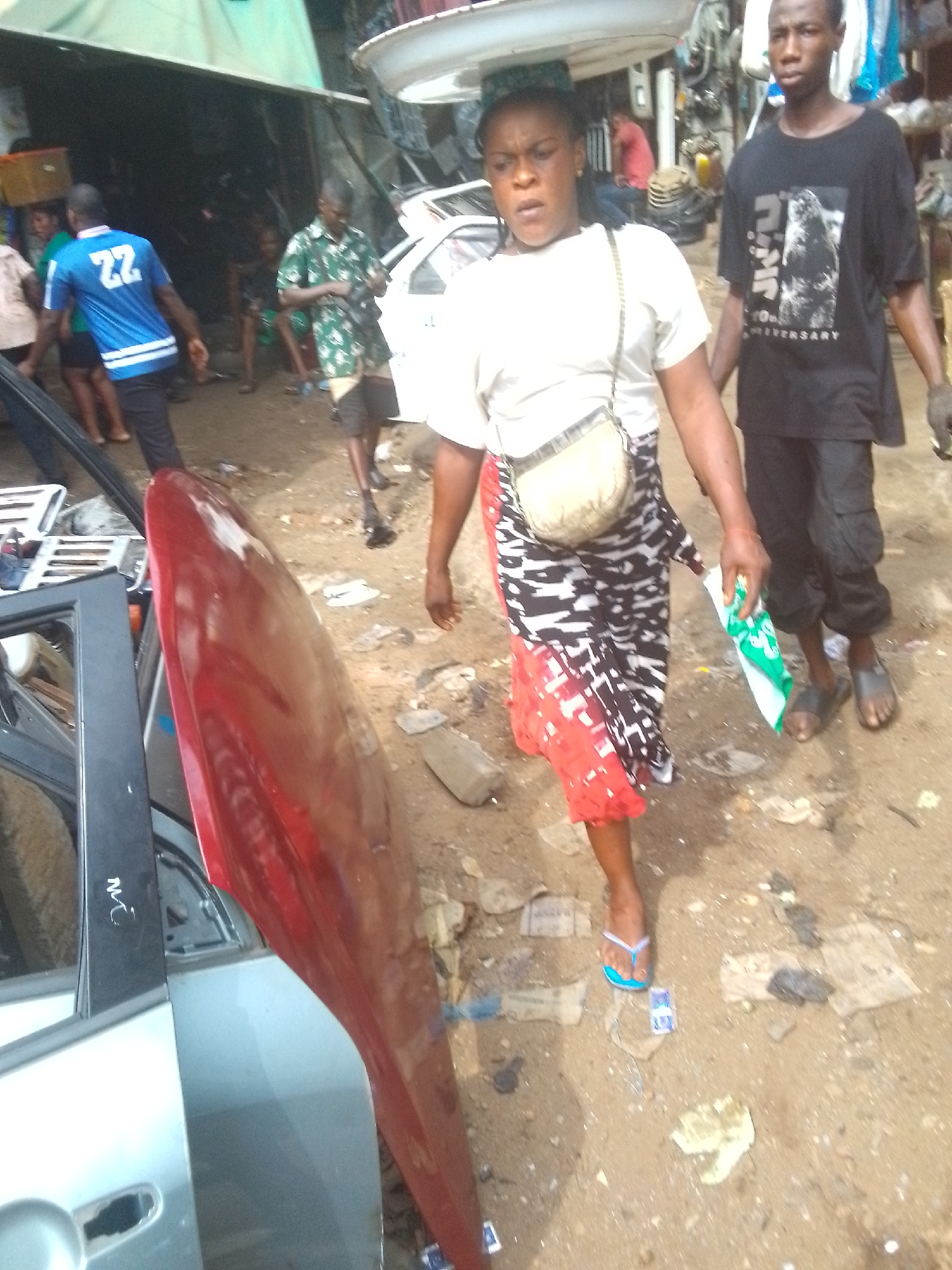 The image captures a vibrant, busy outdoor street or market scene, likely an auto parts market, in Onitsha, Nigeria, under natural daylight. The unpaved ground is dusty and strewn with litter.
In the foreground, a woman walks towards the right, expertly balancing a large, white, round tray or basin on her head. She wears a white t-shirt, a cross-body bag, and a black, white, and red patterned skirt, with light blue flip-flops. She holds a folded green and white item in her right hand, maintaining a neutral expression. Beside her, a young man walks in a black t-shirt featuring a "GODZILLA" graphic and "ANNIVERSARY" text, black cargo trousers, and dark sandals. He has a glove on his left hand, also with a neutral expression.
To the immediate left, a red car hood or fender leans upright, partially revealing a silver car door with "MC" written in white marker on its black frame. Various metallic car parts are visible within the door's opening.
Further back, other people navigate the space. A man wearing a blue t-shirt with the number "22" on the back walks away, while another man in a green patterned shirt walks towards the right. The background is cluttered with makeshift stalls, some covered by tarpaulins, and various auto components, including parts of a white vehicle. The scene conveys the lively, functional atmosphere of a commercial hub.