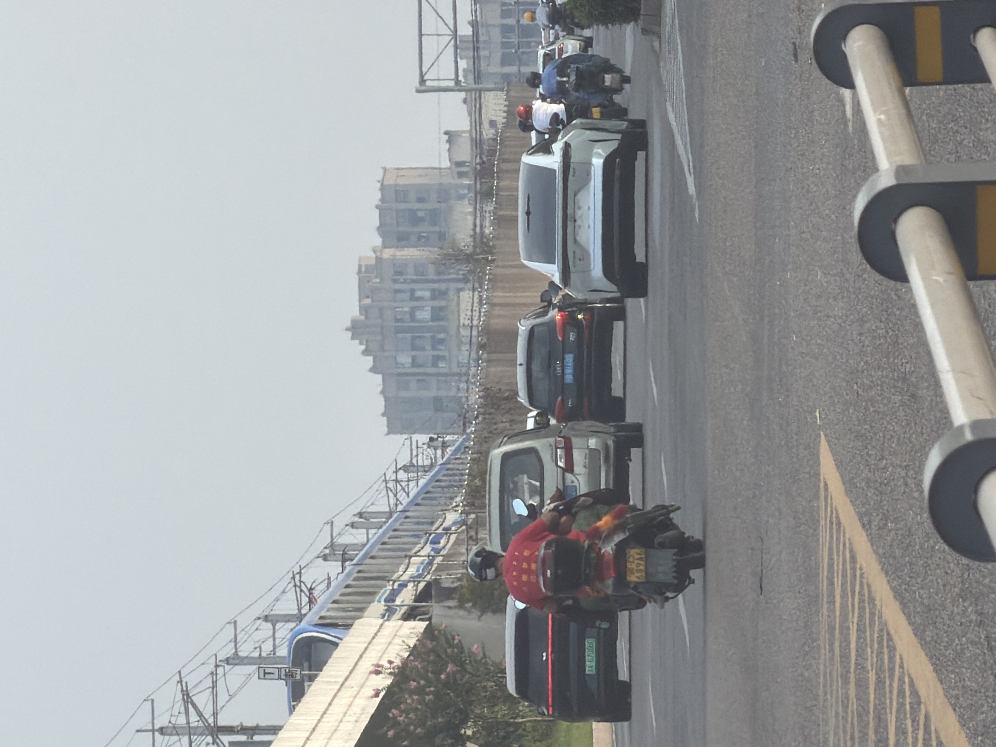 The image captures a street scene in Jiangjin District, Chongqing, China, featuring a traffic-congested road with several vehicles and pedestrians. In the foreground, a motorcycle with a rider wearing a red shirt and a helmet is visible, followed by a silver van. To the left, a black SUV with license plate "AA 67066" is parked. Further ahead, a white car with a distinct design is traveling in the same direction, with two motorcycles behind it. In the background, a metro train is seen on an elevated track, alongside a multi-story building with a facade suggestive of older architecture. The environment appears to be an urban setting with clear skies.