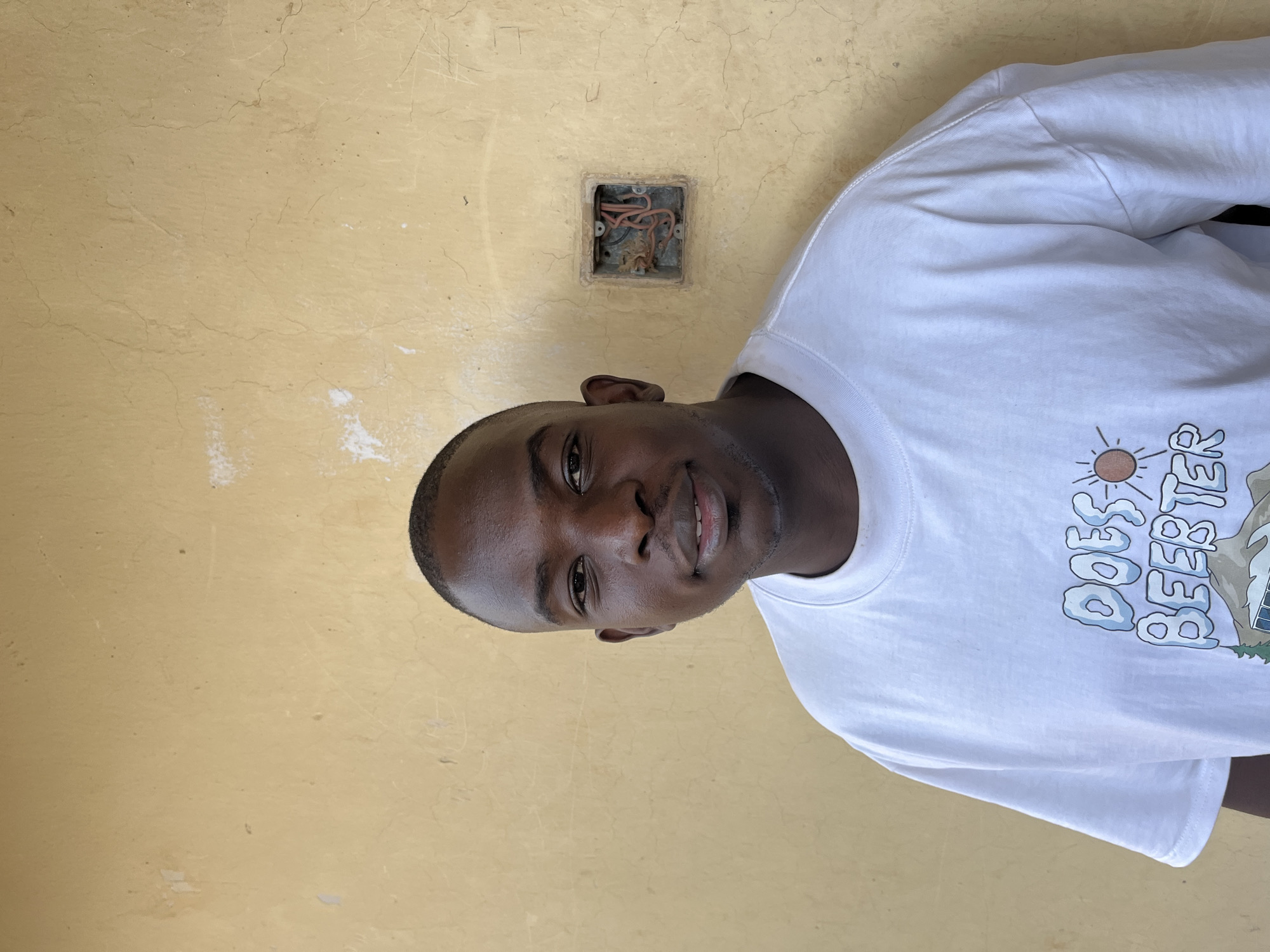 A young Black man with short hair and a light beard smiles directly at the camera. He is wearing a white t-shirt with a graphic on the front that says "DOES BEERTER" with stylized letters. The man is standing in front of a textured, light yellow wall with some cracks and a disused electrical outlet visible on the right side of the frame. The scene appears to be outdoors, possibly in a residential or commercial area in Jos, Nigeria, given the architectural style of the wall and the presence of exposed electrical wiring, which is common in some parts of the city.