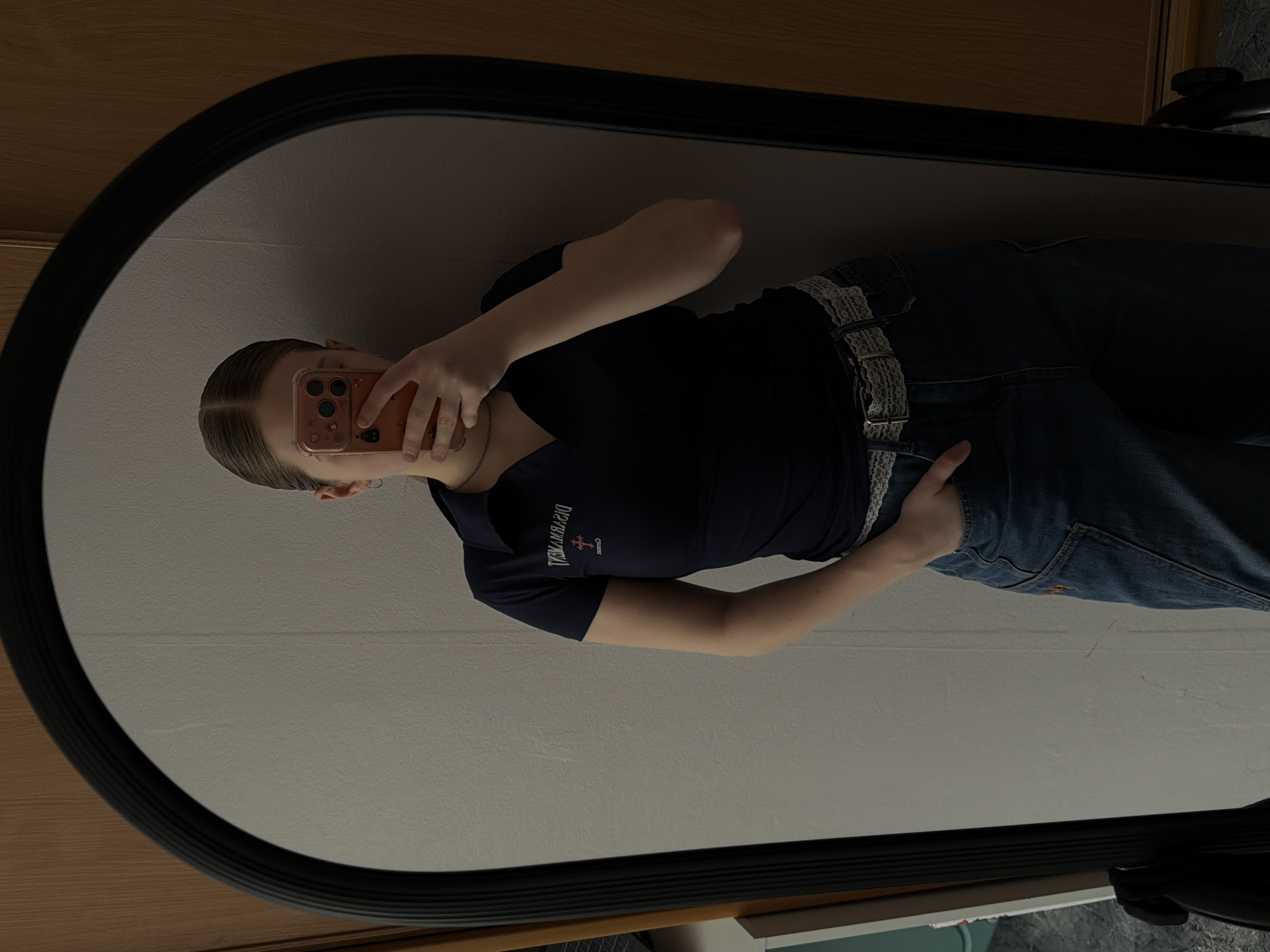 A person is taking a mirror selfie with a phone. The person is wearing a dark blue shirt with white lettering, blue jeans with a lace belt, and a necklace. The mirror has a thick black frame. The background appears to be a wall in a room. There is a wooden surface visible above the mirror. The city of Perm, Russia, is not discernible from the image.