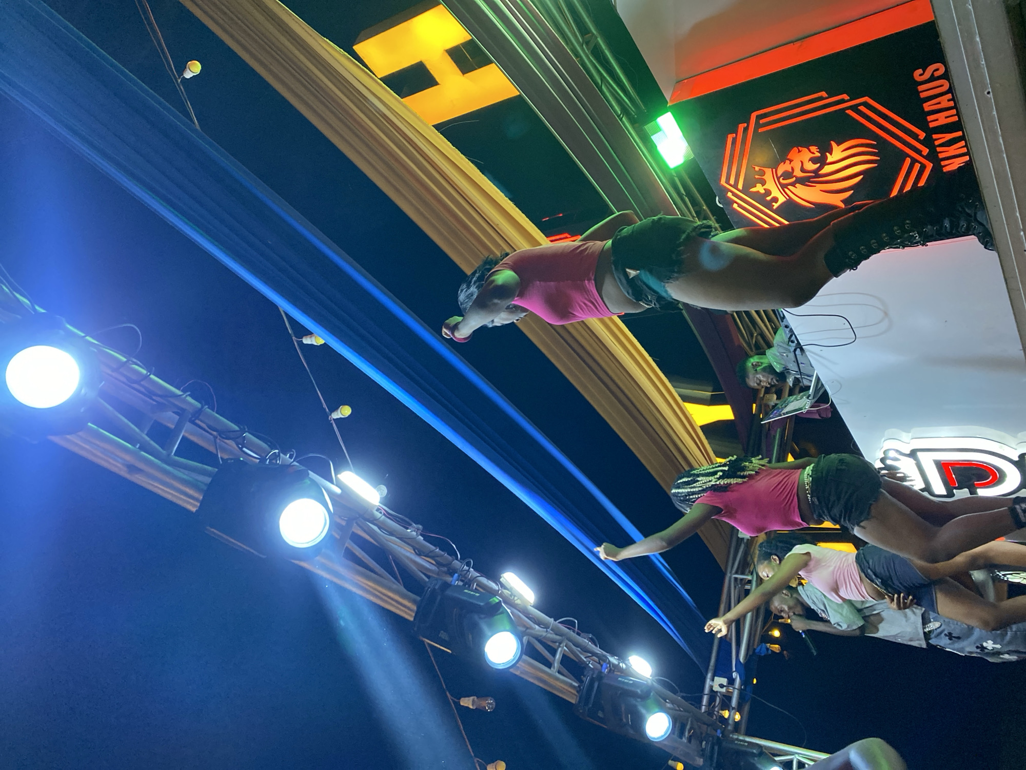 The image depicts a nighttime outdoor performance on a stage in Gura Top, Nigeria. A group of individuals are on stage, with a DJ set up in the background. Two women in pink tops and dark shorts are dancing, one with her arm raised and the other with her hand on her hip. Another woman in a pink top and dark shorts is also dancing. A man in a light-colored shirt and patterned pants is holding a microphone and appears to be singing or speaking. The stage is decorated with fabric draped across the top, and there are various lights illuminating the scene. A large, illuminated logo featuring a lion's head and the text "UNKY HAUS" is visible on the right side of the stage. The overall atmosphere suggests a lively event.