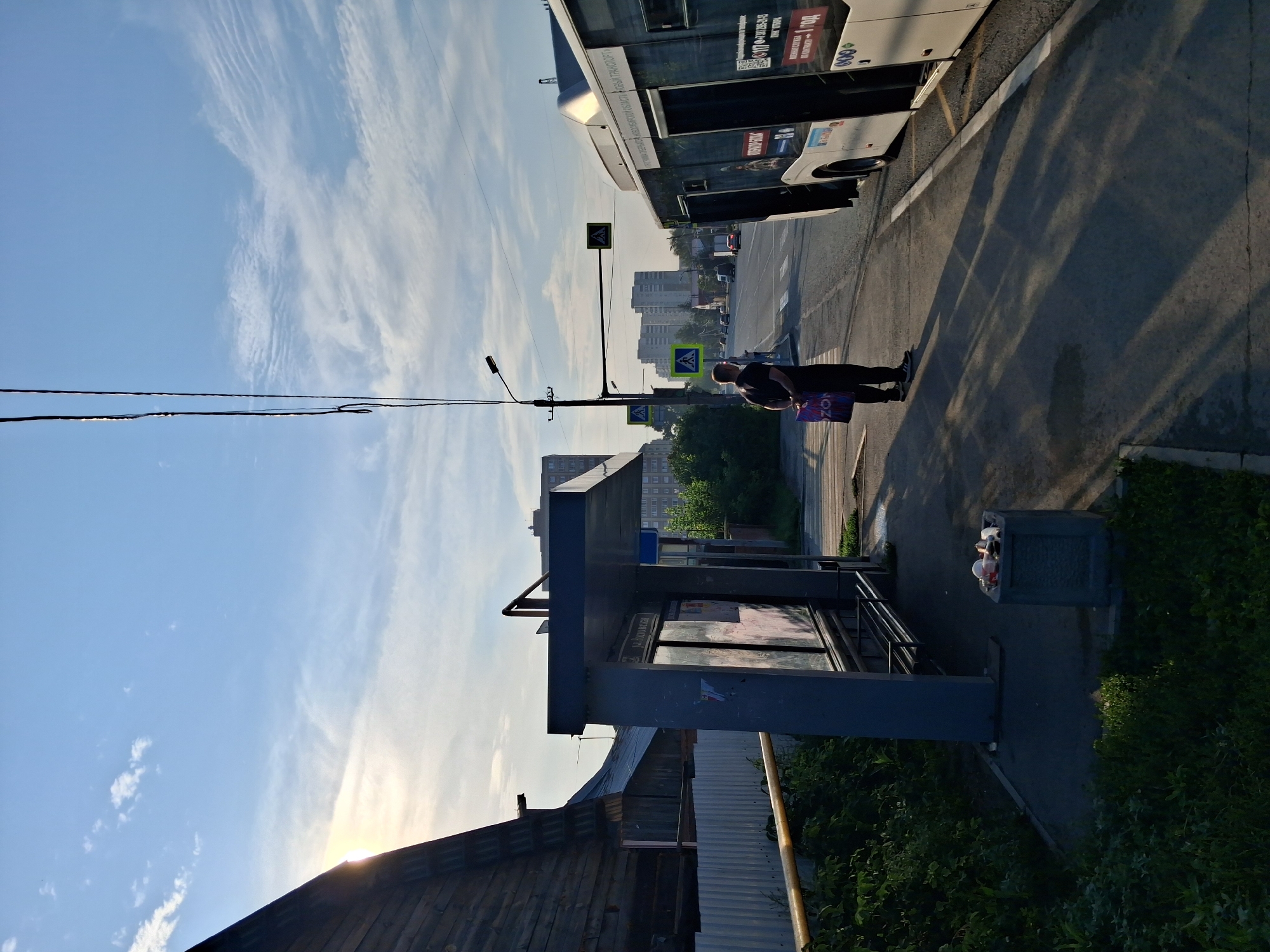 A bus stop is located on a city street in Novosibirsk, Russia, under a bright, partly cloudy sky. A man in a black t-shirt and dark pants stands on the sidewalk, holding a shopping bag. A white bus with its doors open is parked at the bus stop. A bus shelter is to the left of the man, and a trash can is visible near the shelter. In the background, there are residential buildings, trees, and traffic on the street. The sun is low in the sky, casting long shadows across the sidewalk.