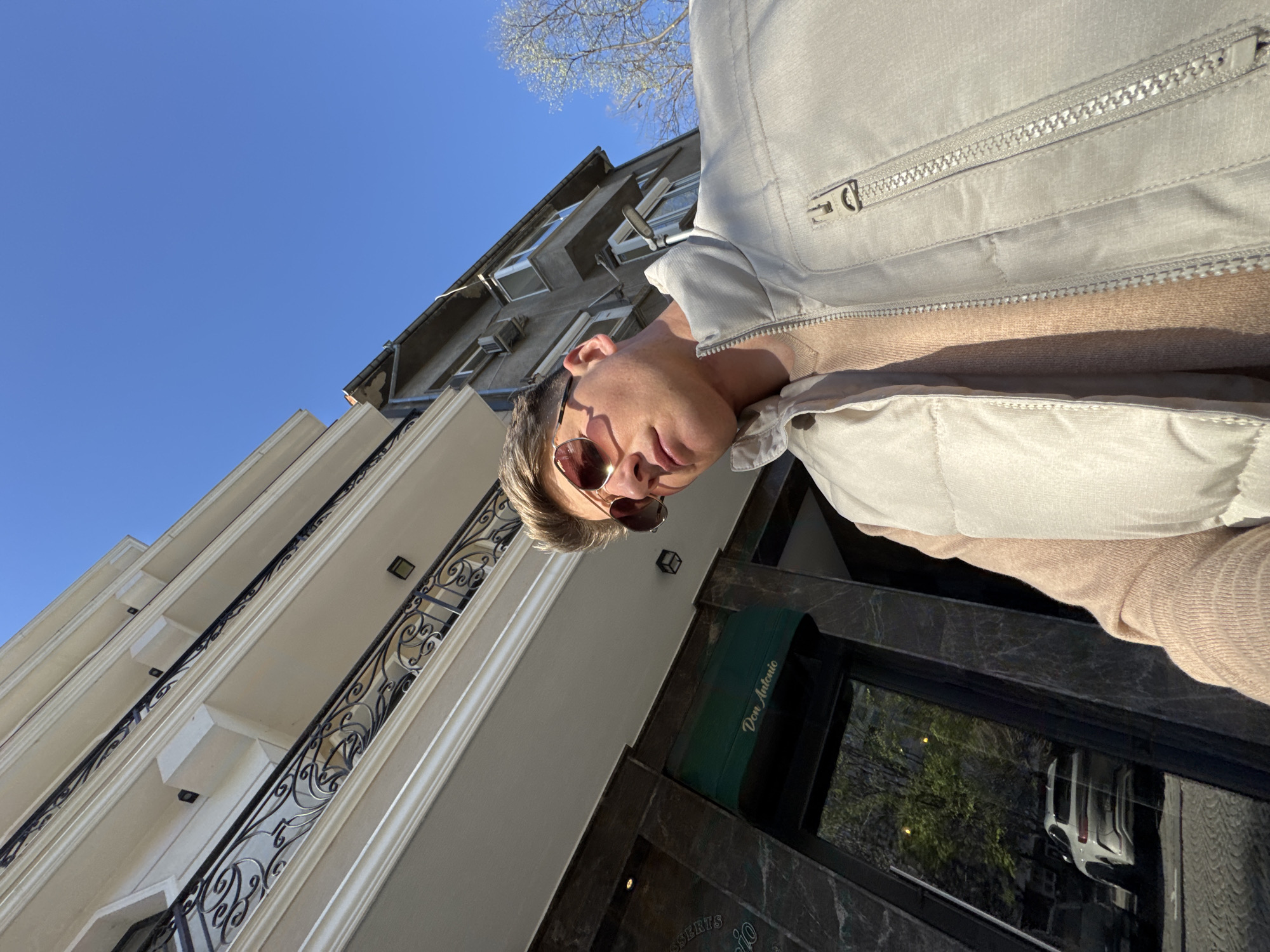 The image shows a person wearing sunglasses and a light-colored jacket standing in front of a building with a dark marble facade. A green awning with the words "Don Antonio" is visible above the entrance. The building has multiple floors with balconies featuring ornate black iron railings. A reflection of a white van can be seen in the window. The sky is bright blue. The location is likely in Plovdiv, Bulgaria, based on the awning inscription "Don Antonio".