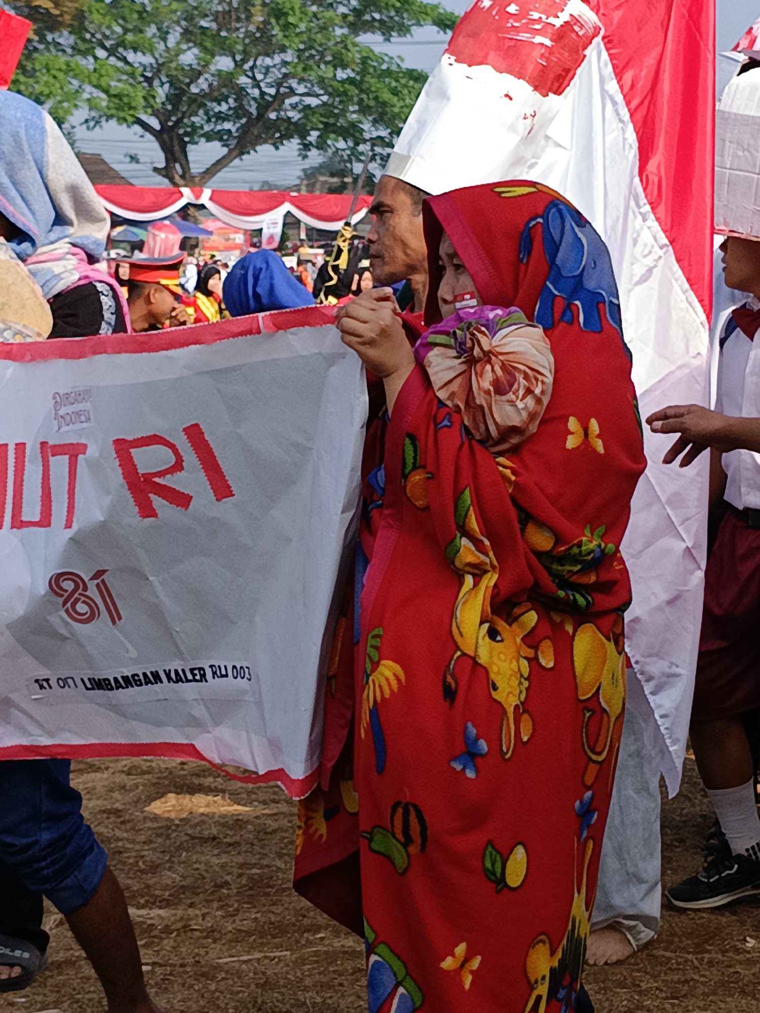 This is a close-up, eye-level, outdoor shot showing a group of people participating in a parade or public event. The foreground is dominated by a white banner with red text, partially obscured by individuals. The banner reads "N UT RI" in large red letters, with smaller text below that says "RT 011 LIMBANGAN KALER RLJ 003". A red fabric with a colorful animal and butterfly print covers the right side of the frame, worn by a person whose face is partially visible. Another person, a man, is visible to the left of the banner, looking down and to the side. In the background, a large Indonesian flag, red and white, is prominently displayed, along with other flags and colorful decorations, suggesting a celebratory atmosphere. A tree with green foliage is visible in the upper left quadrant. The ground appears to be covered in dry, light-colored grass or straw. The lighting indicates it is daytime, with a bright sky. The event appears to be taking place in Cibodas, Indonesia, as suggested by the text on the banner.